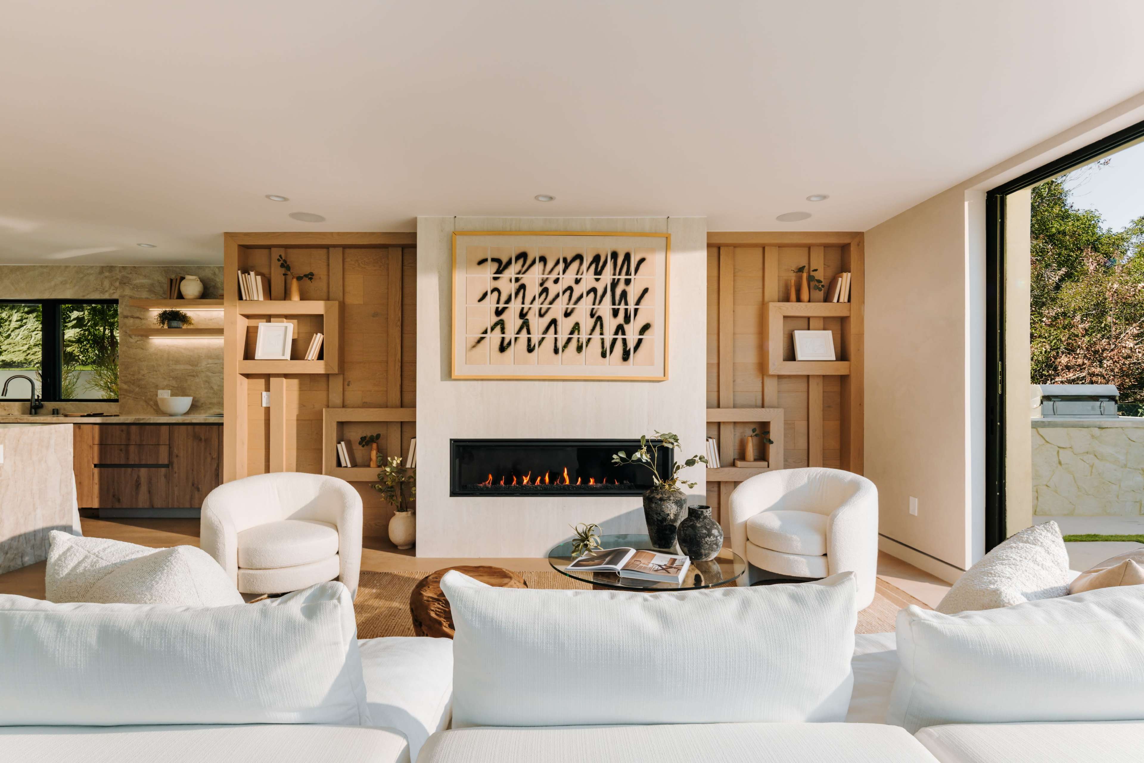 The image shows a modern living room featuring a minimalist design with two white armchairs, a coffee table, and a wall-mounted fireplace beneath an artistic framed print.