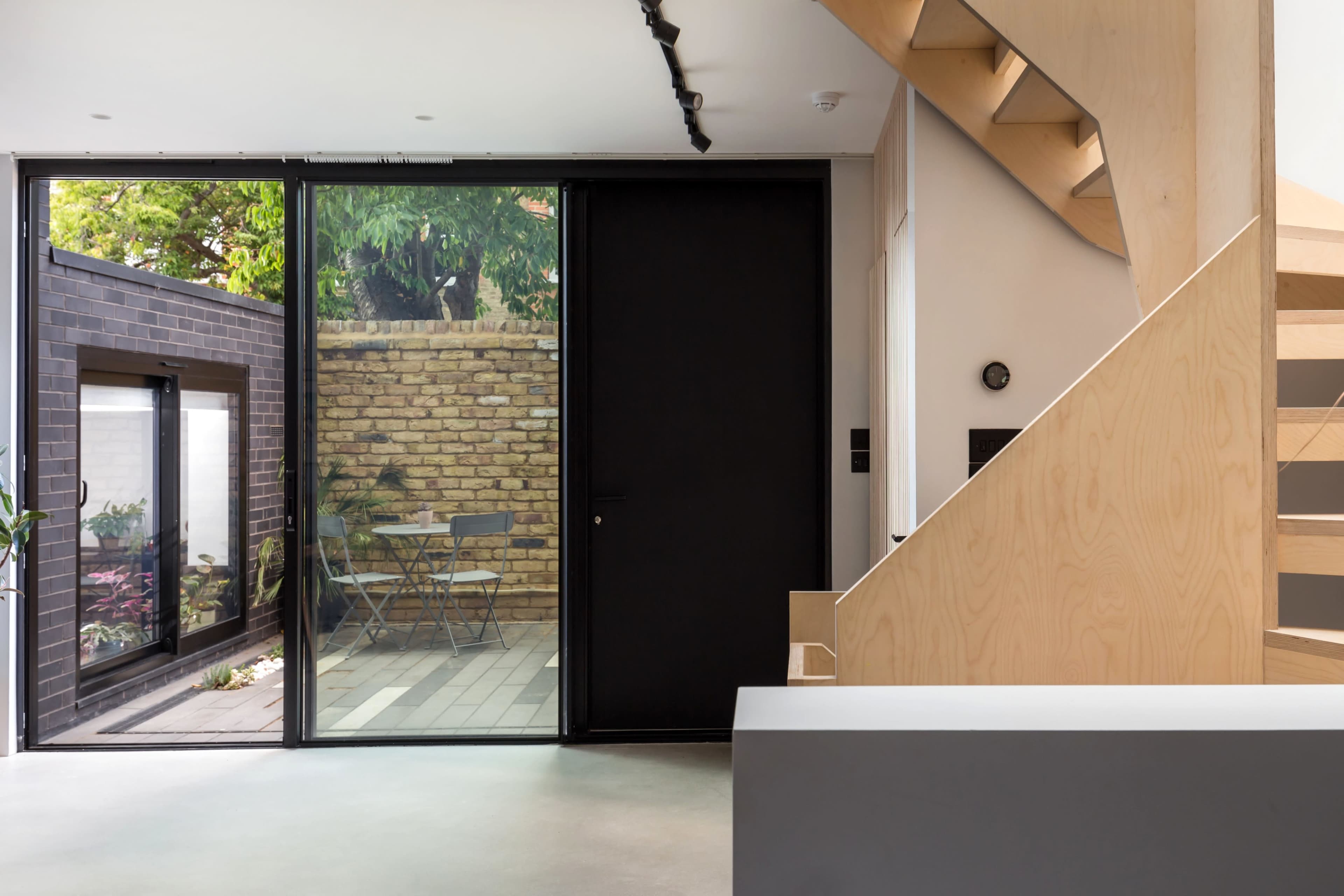 A modern indoor space with a staircase leading up, a large glass door opening to a brick courtyard with outdoor seating, and a wooden panel accent.