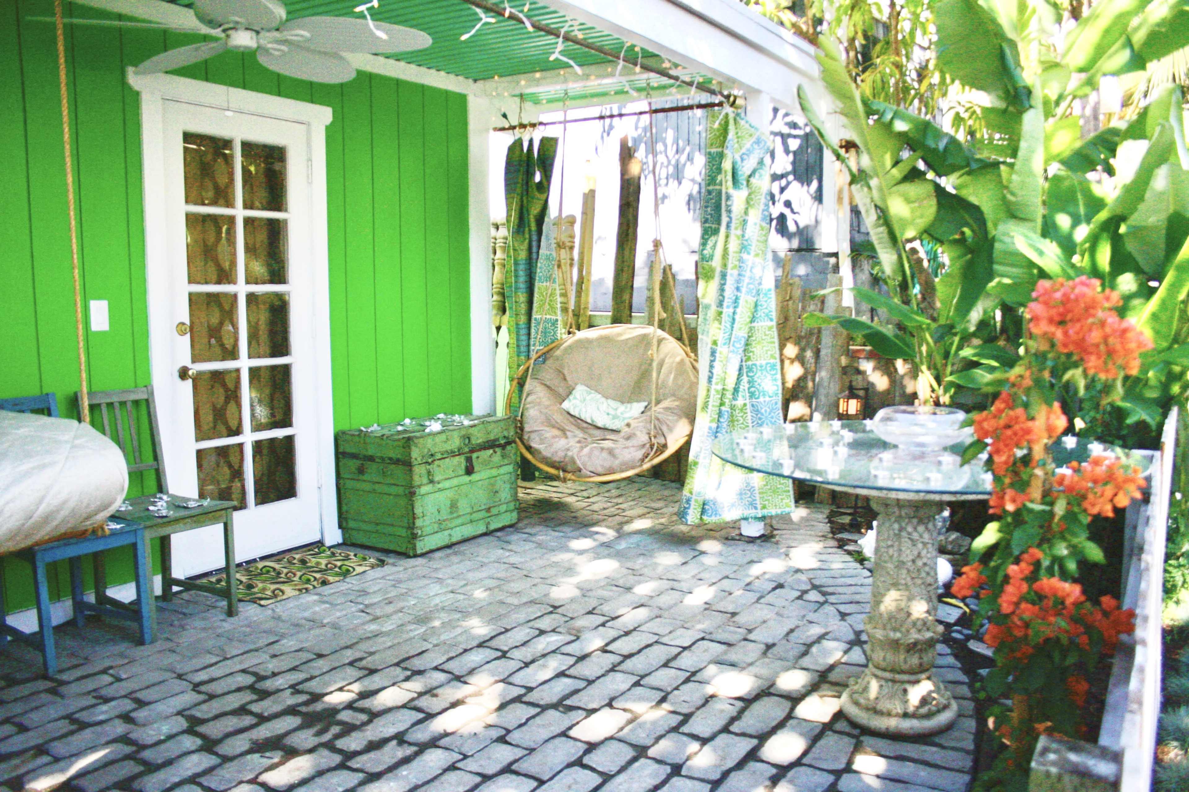 The image features a bright green outdoor patio with a round glass table, a cushioned chair, and a large hanging chair surrounded by tropical plants.
