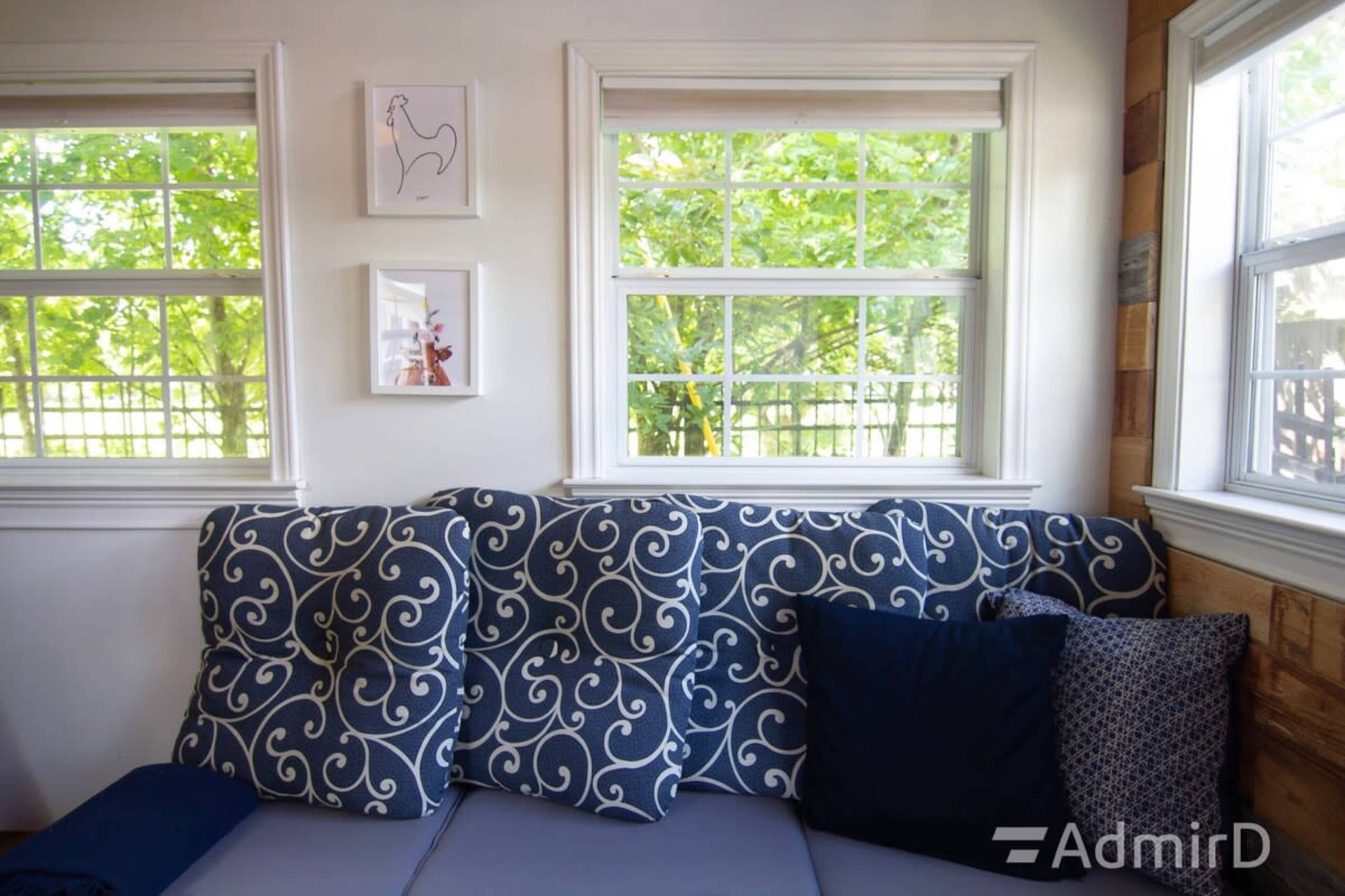 The image shows a corner seating area with a patterned upholstered backrest and decorative pillows, flanked by two windows that overlook greenery outside.