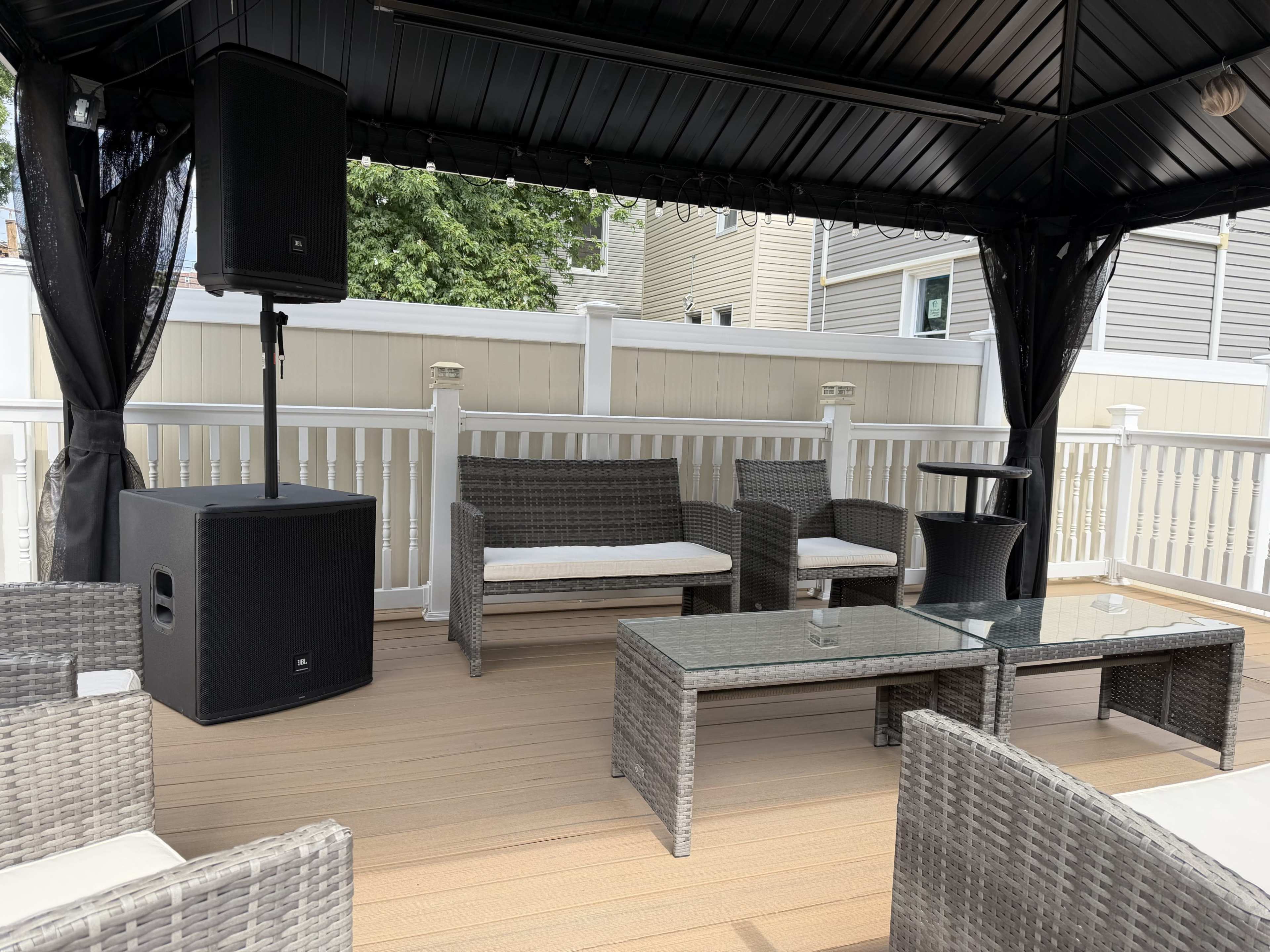 The image shows a covered outdoor seating area with wicker furniture, a glass coffee table, and a large speaker set up in one corner.