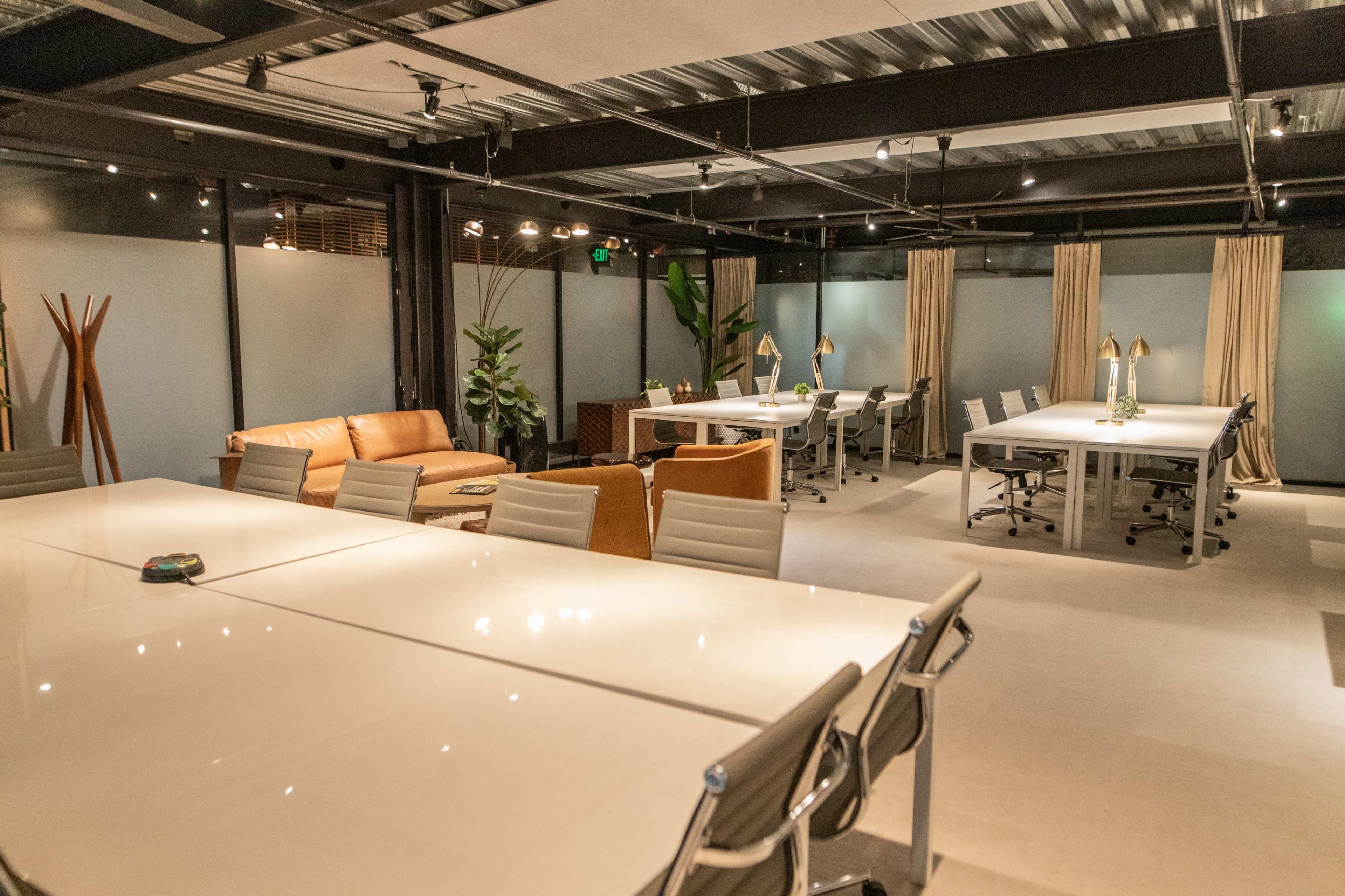 The image shows a modern office space with multiple tables, chairs, and a lounge area featuring two sofas and potted plants.