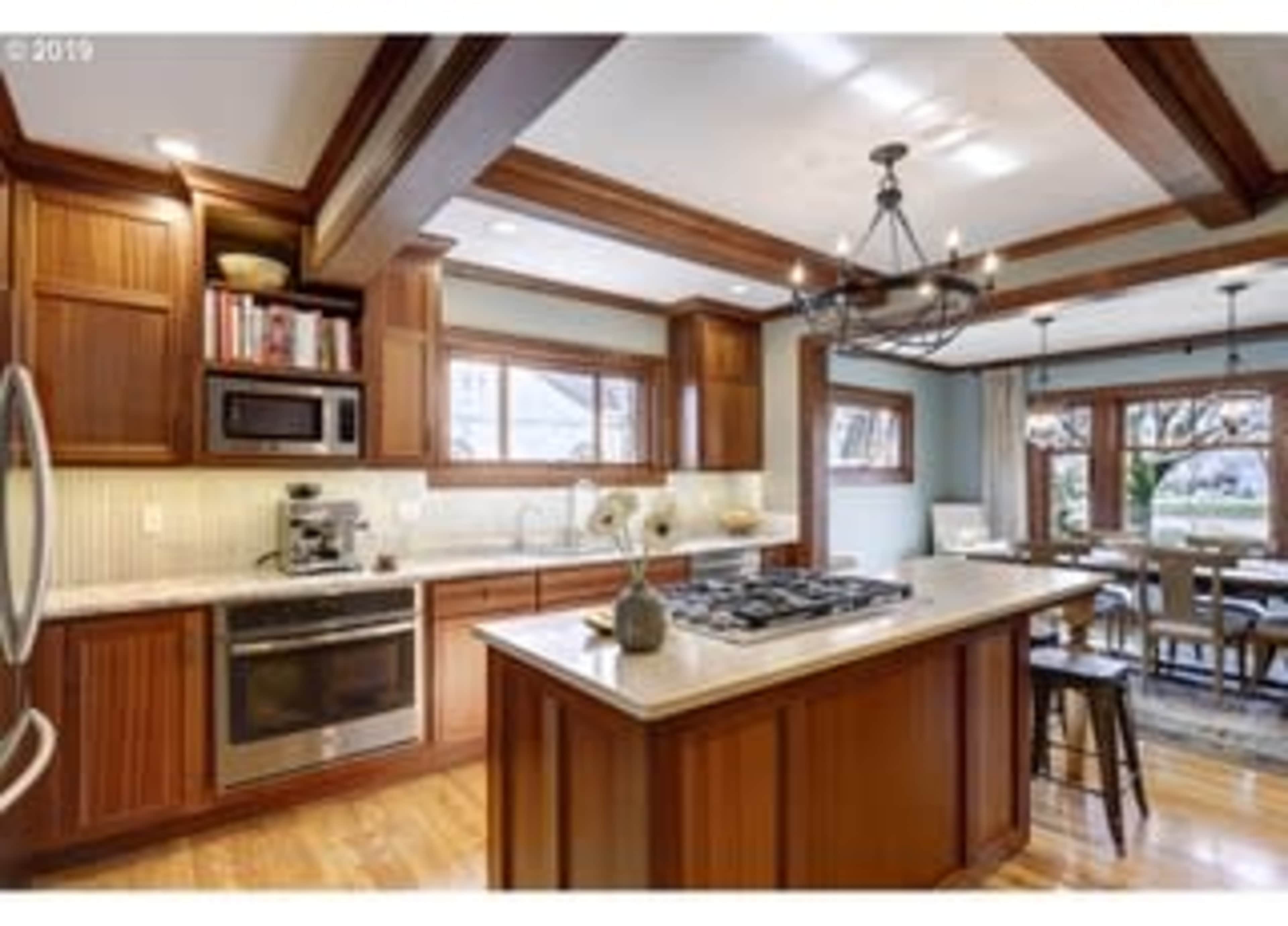 A modern kitchen features wooden cabinetry, stainless steel appliances, a large island with a gas range, and a dining area with large windows.