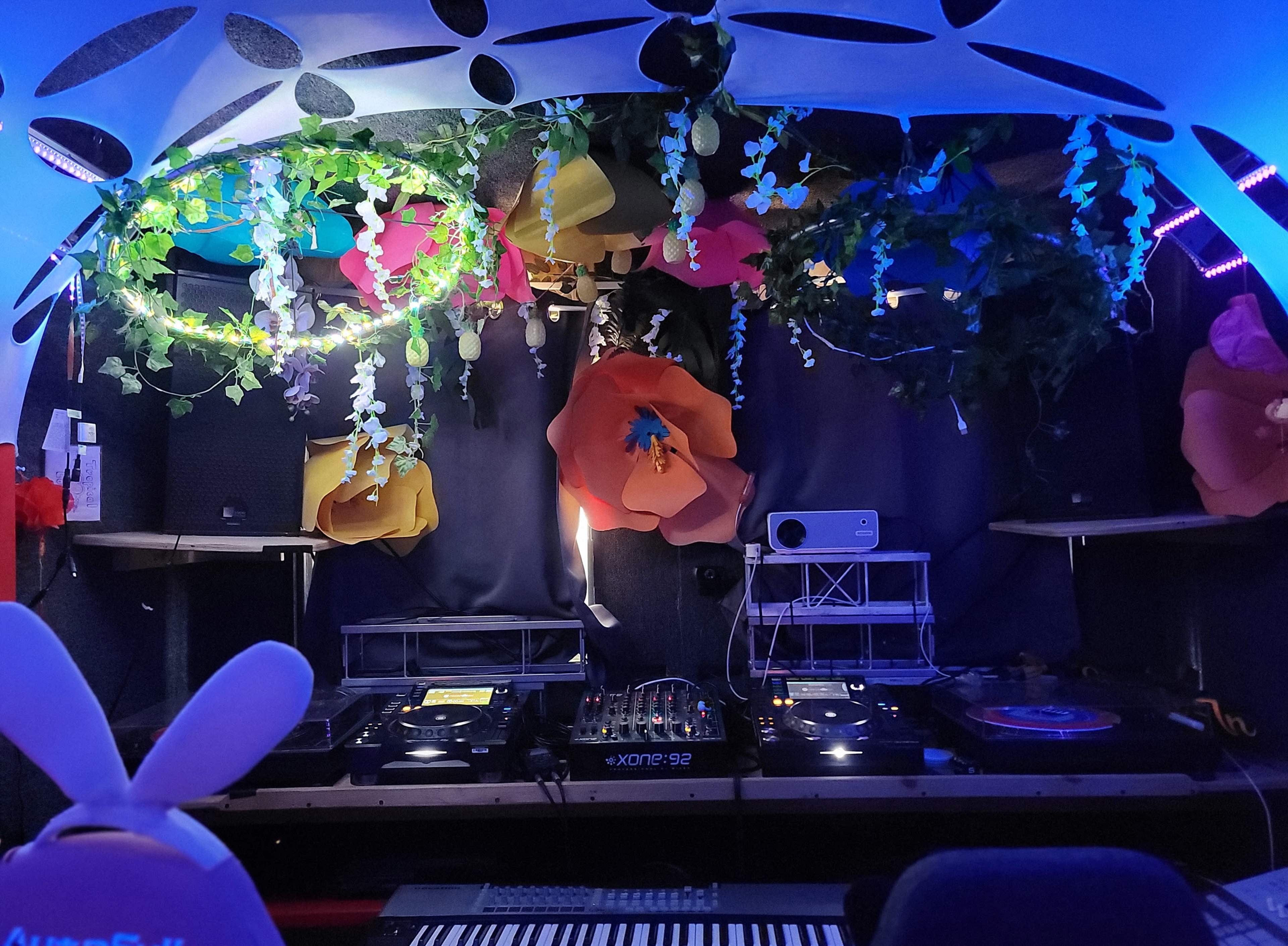 A DJ booth is set up in a decorated space featuring colorful flowers, greenery, and sound equipment.