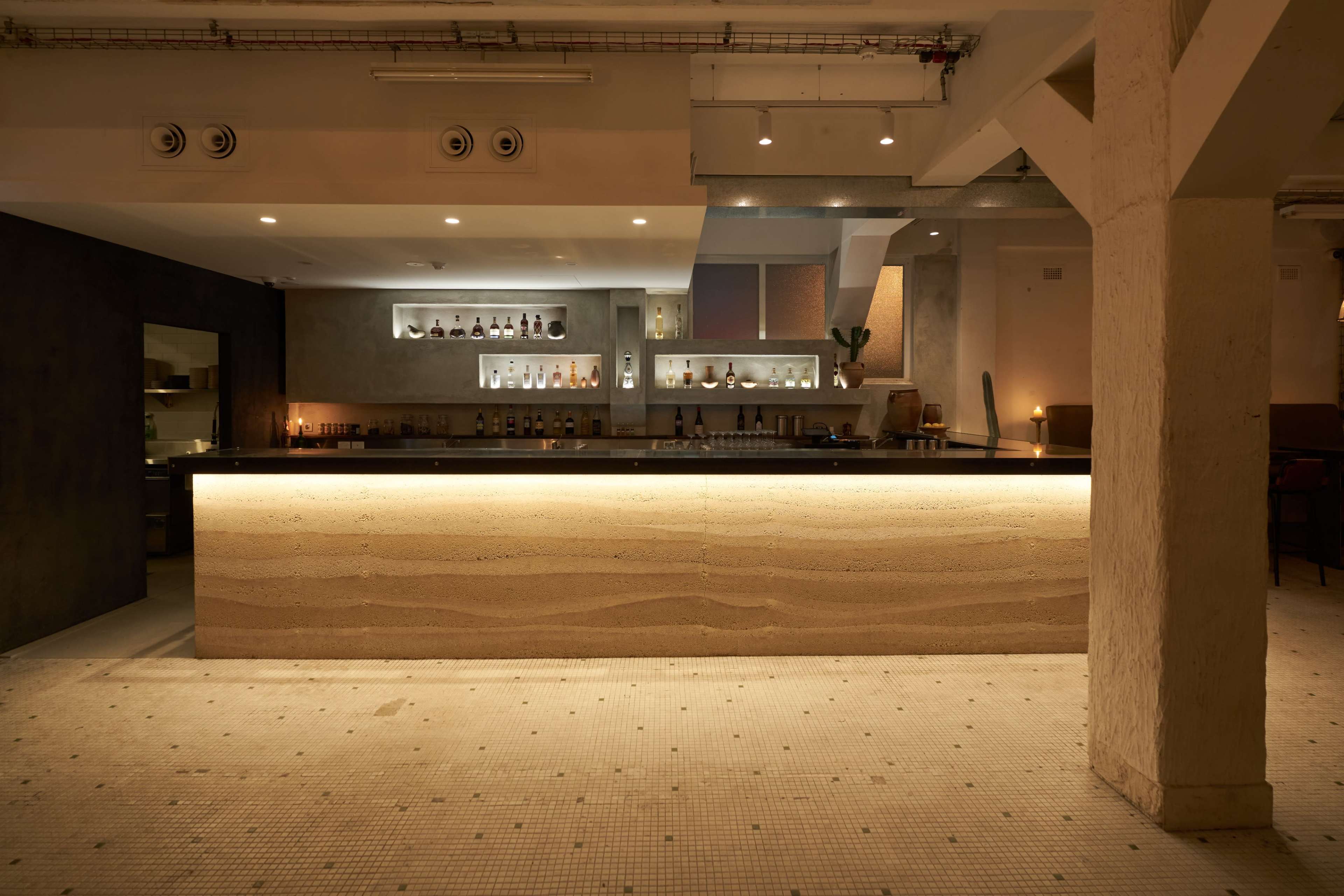 The image shows a softly lit bar area with a minimalist design featuring a long counter made of light-colored material and shelves displaying various bottles in the background.