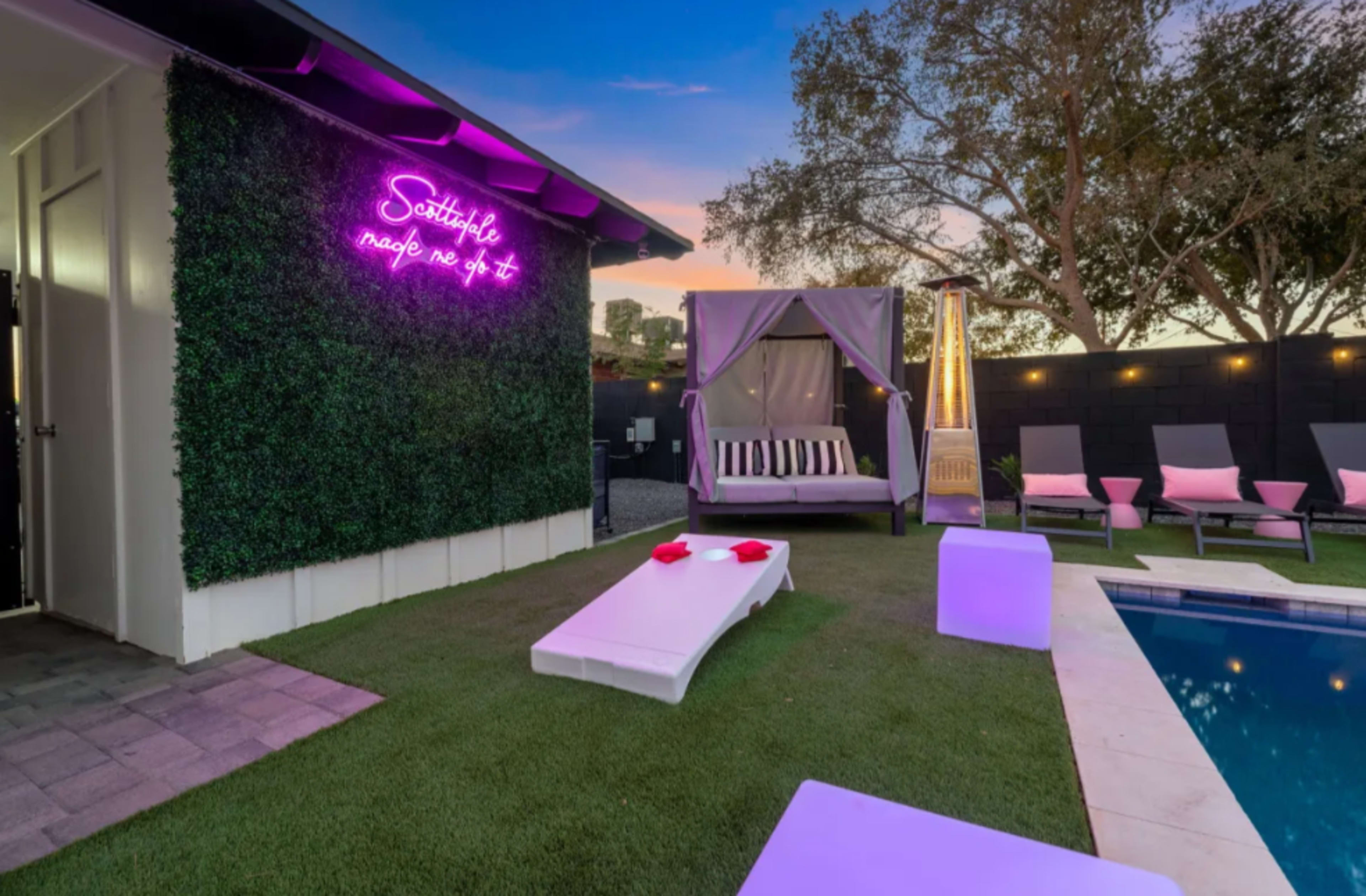 A modern outdoor space features a swimming pool, a lounging area with pink chairs, a canopy bed, and neon signage on a green wall.