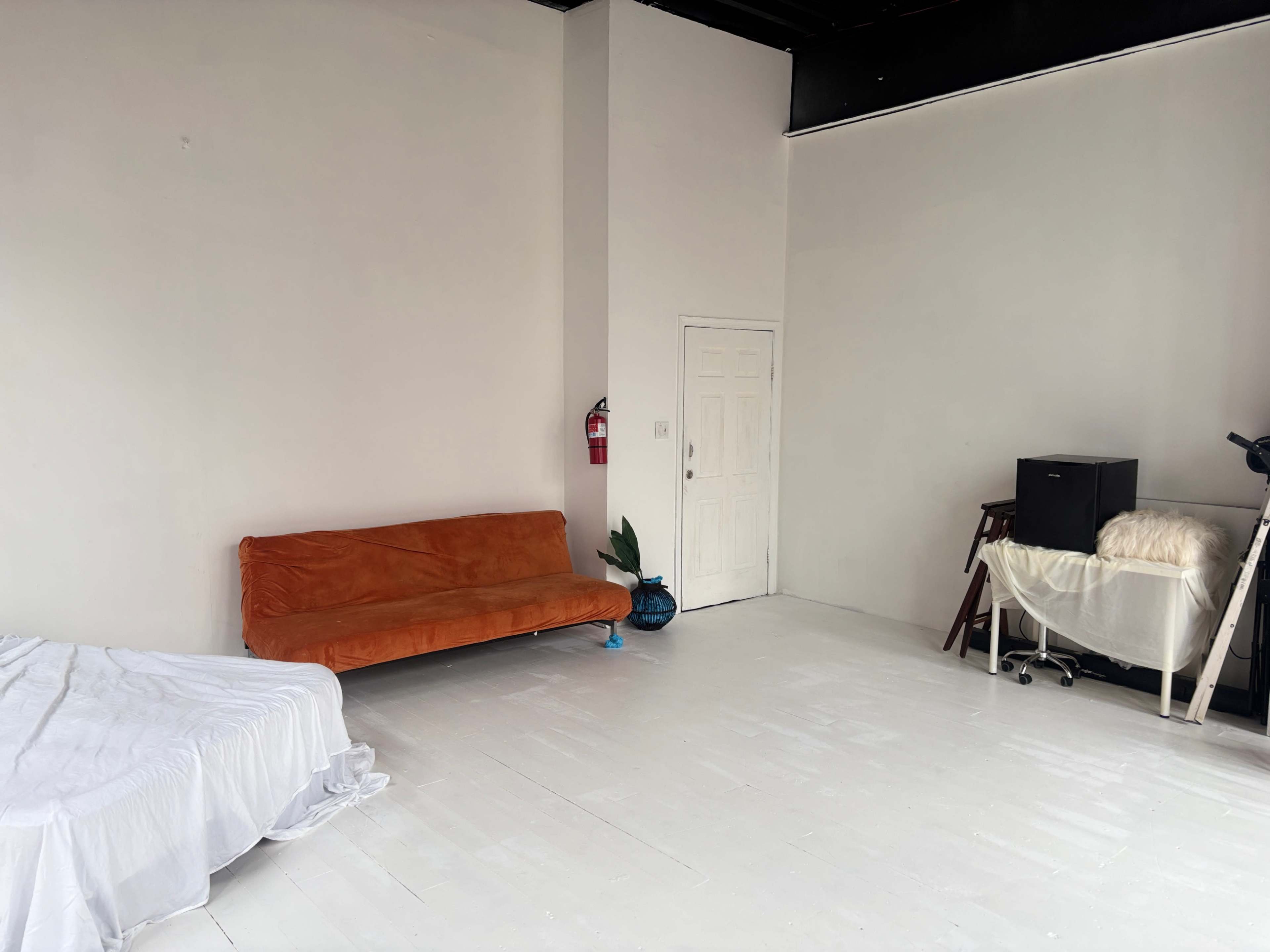 The image shows a spacious, minimalist room with an orange couch, a white bed with a sheet, a blue decorative vase, and a small desk with a chair.