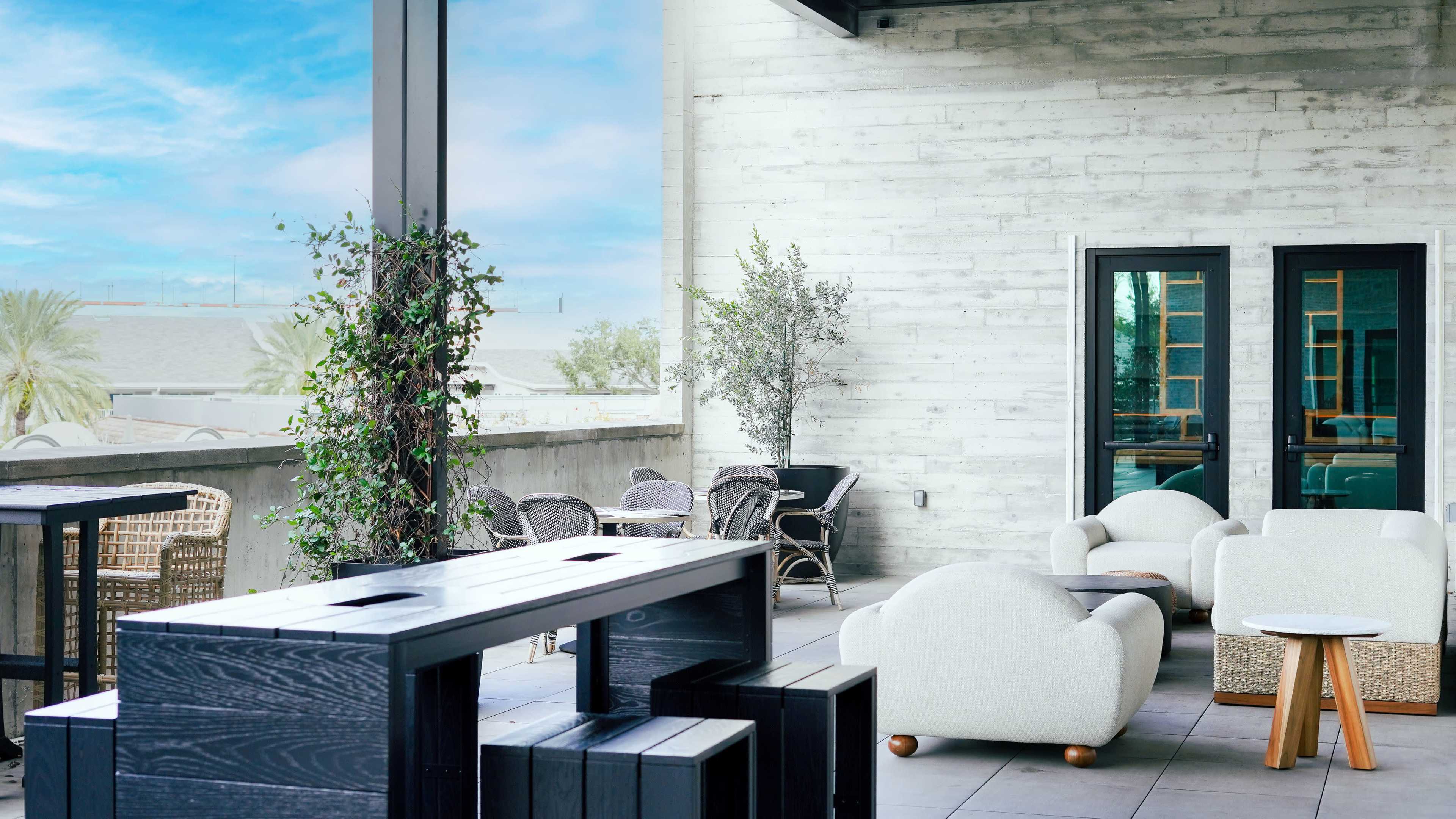 The image shows a modern outdoor seating area with white cushioned chairs and a wooden bar counter, surrounded by greenery and large windows.