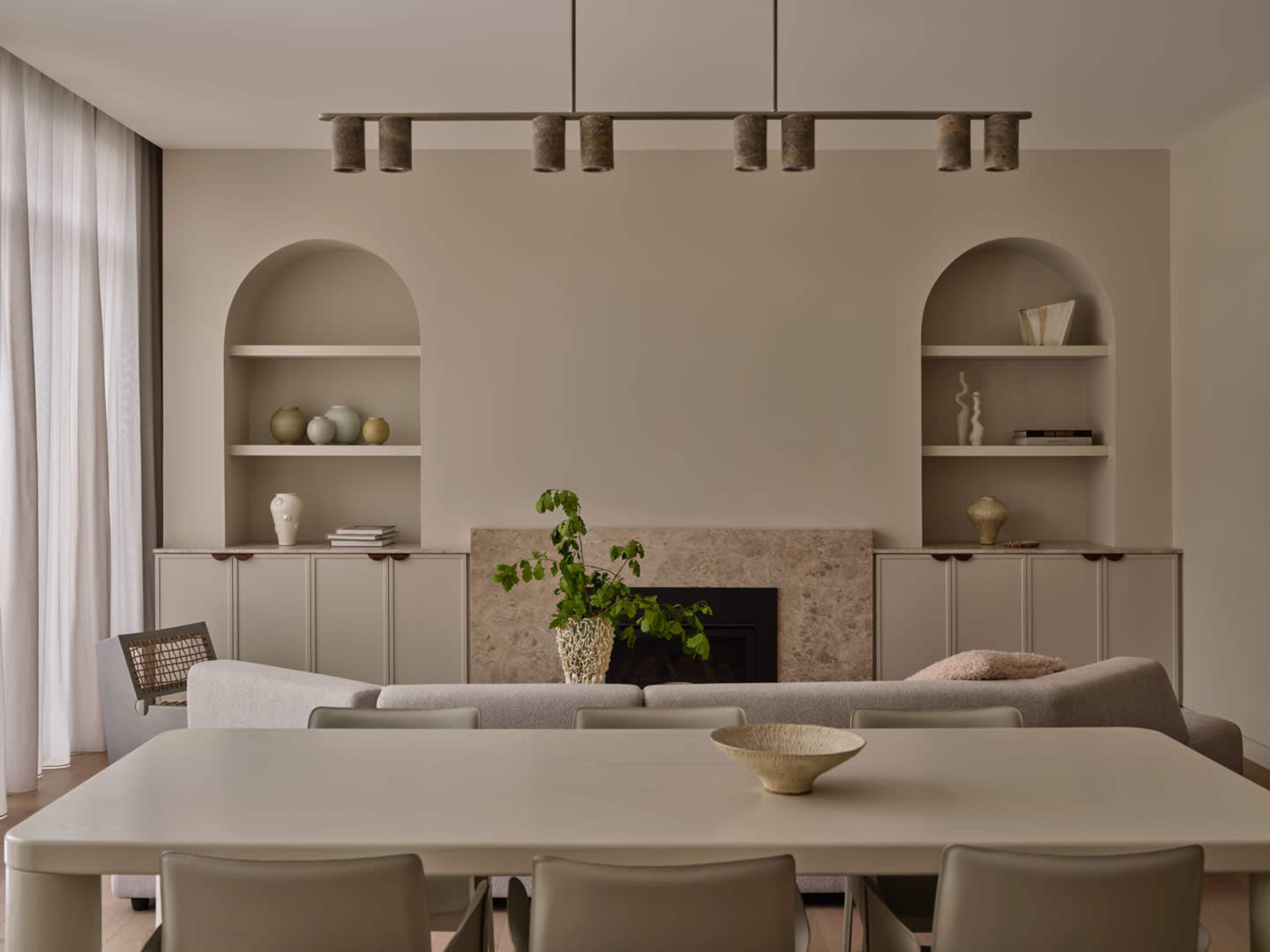 A modern living room features a light-colored sofa, a minimalist dining table, and shelves with decorative objects, set against a neutral wall and large windows with sheer curtains.