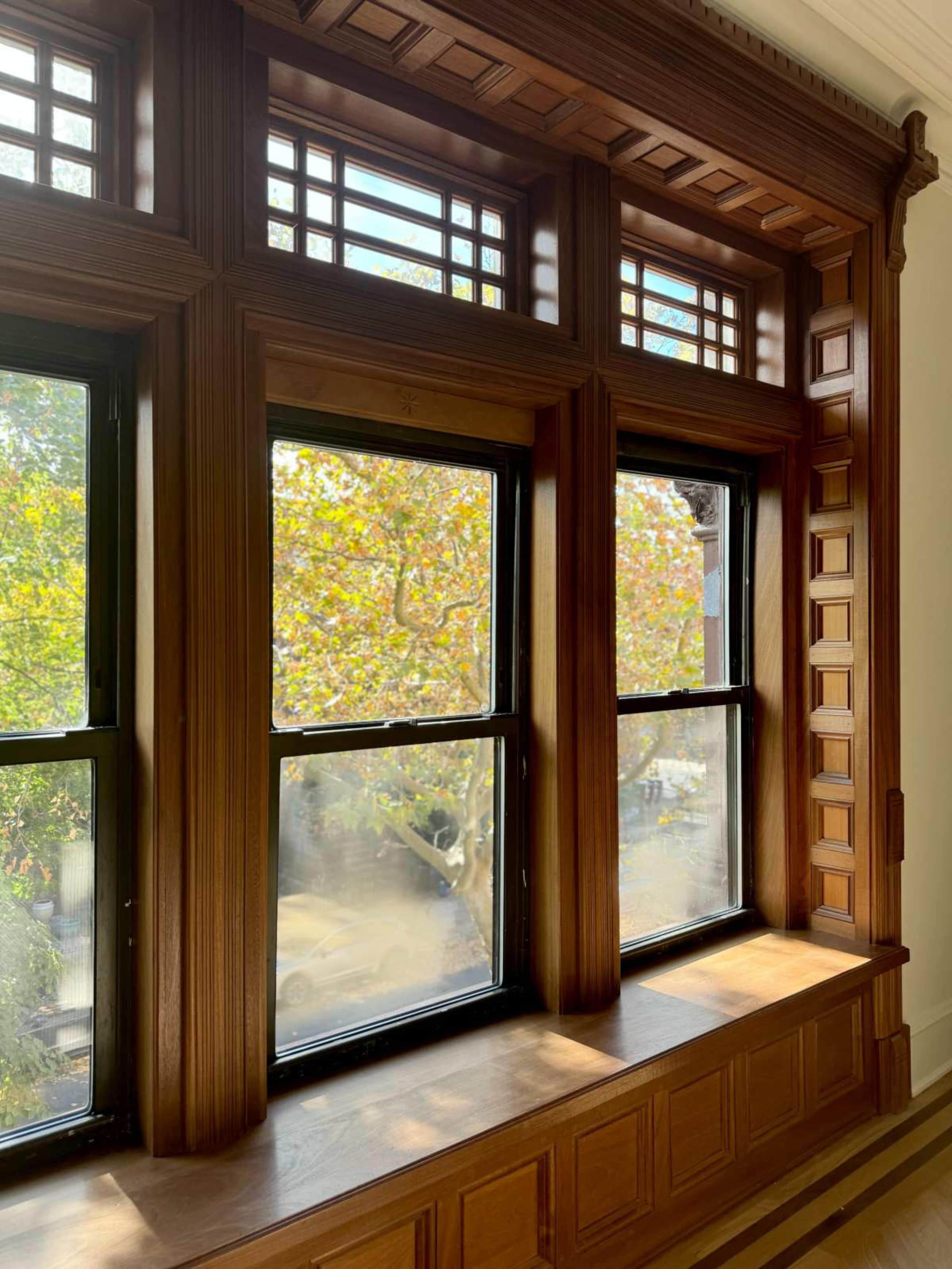 The image shows a decorative wooden window with three panes, framed by intricate woodwork and offering a view of trees outside.