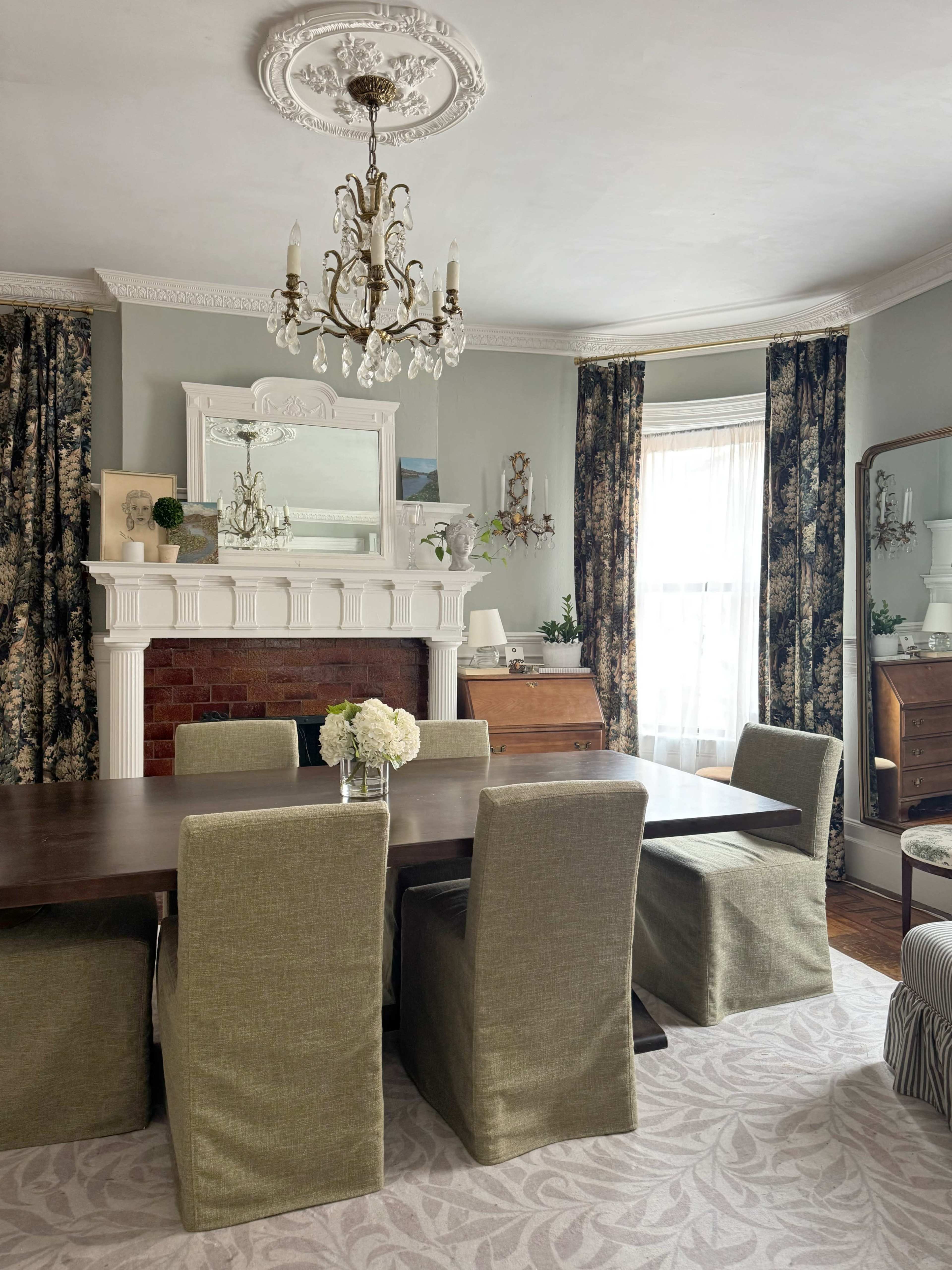 The image shows a dining room featuring a wooden table surrounded by upholstered chairs, with a fireplace adorned with a mirror and decorative elements, and large windows dressed with floral curtains.