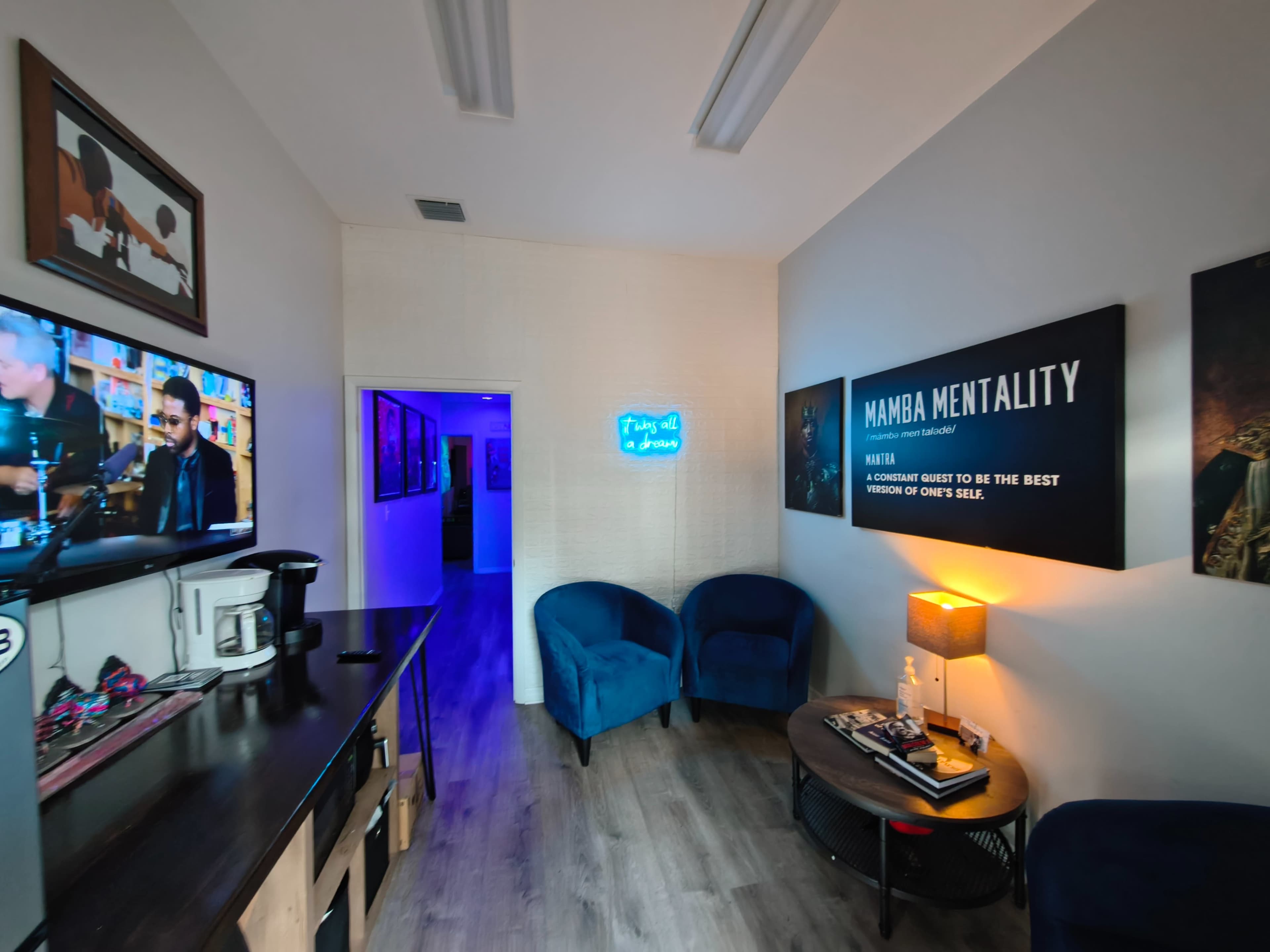 The image shows a small, modern waiting area with two blue chairs, a coffee station, a television playing a show, and walls decorated with artwork and inspirational quotes.