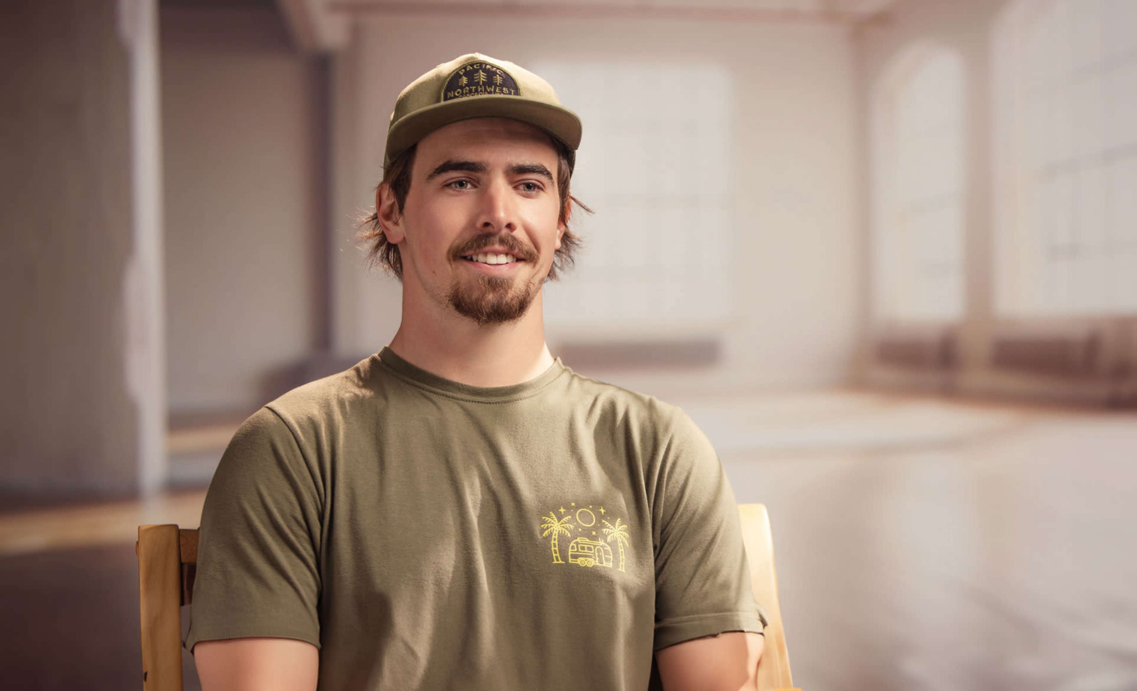 A man wearing a green cap and shirt sits on a chair in a brightly lit room with a wooden floor.