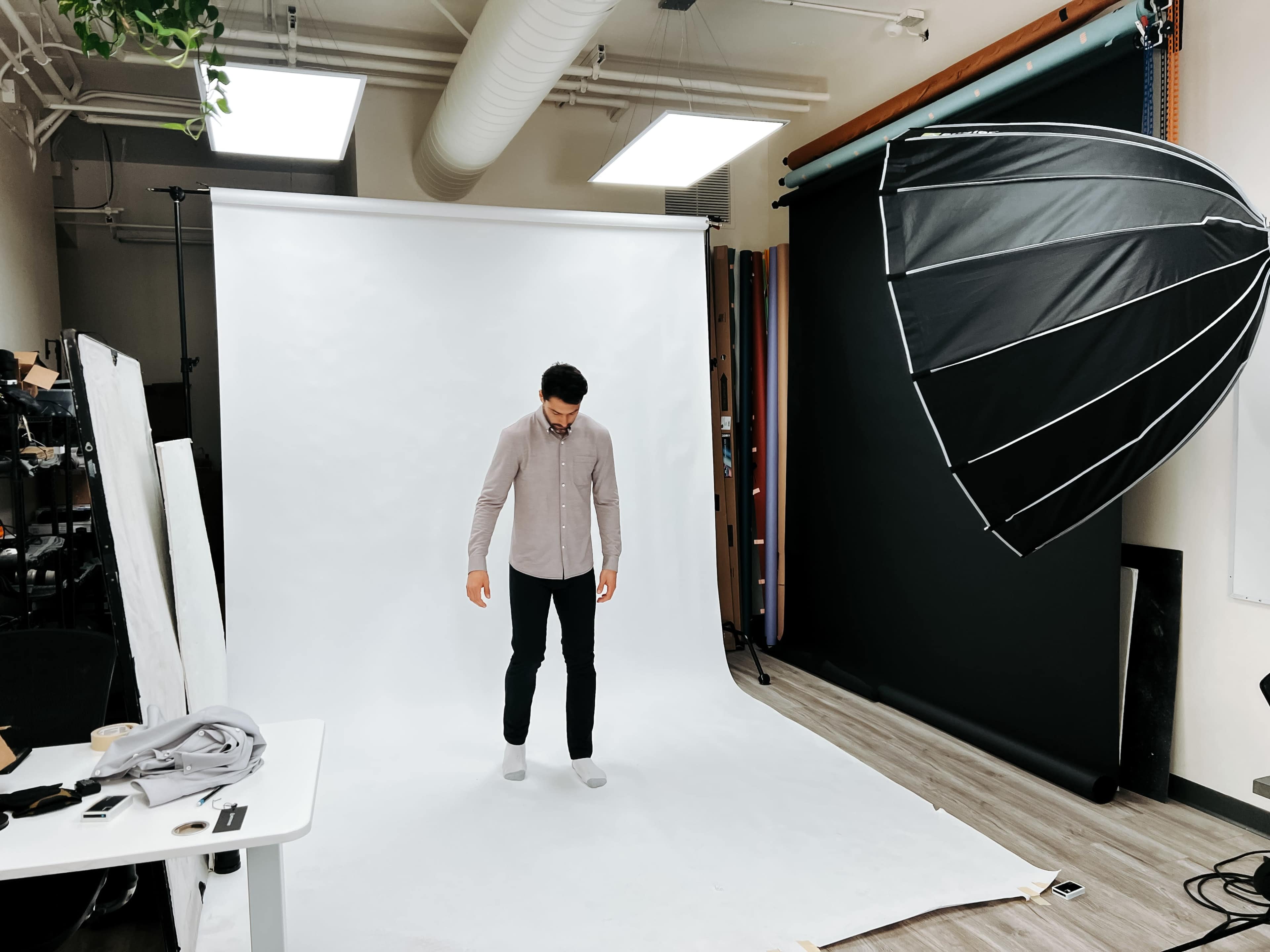 A person stands on a light-colored backdrop in a photography studio, with an umbrella light positioned to the side.