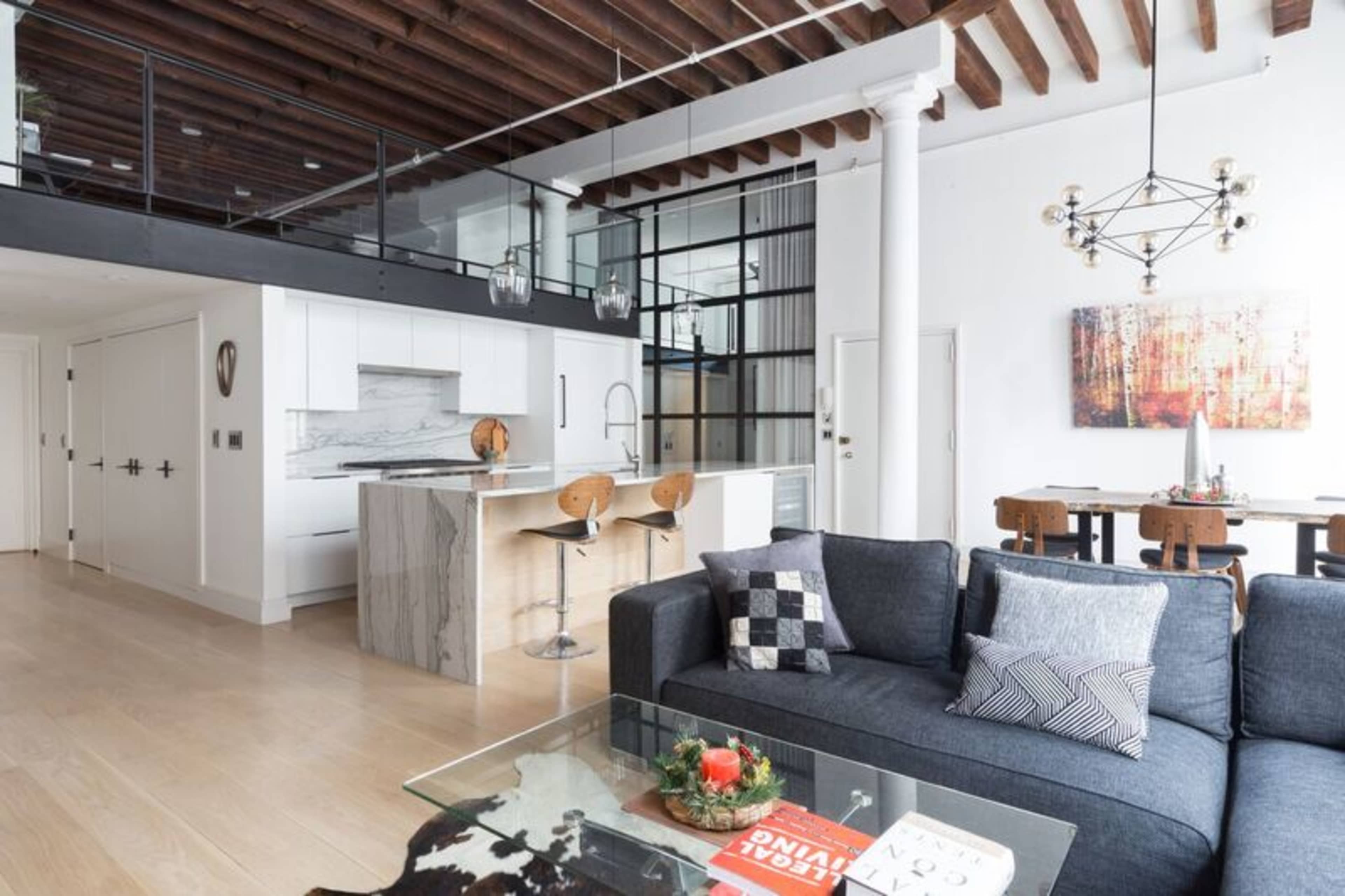A modern open-concept living space featuring a kitchen with a marble island, a seating area with a dark sofa, and a dining table with wooden chairs.