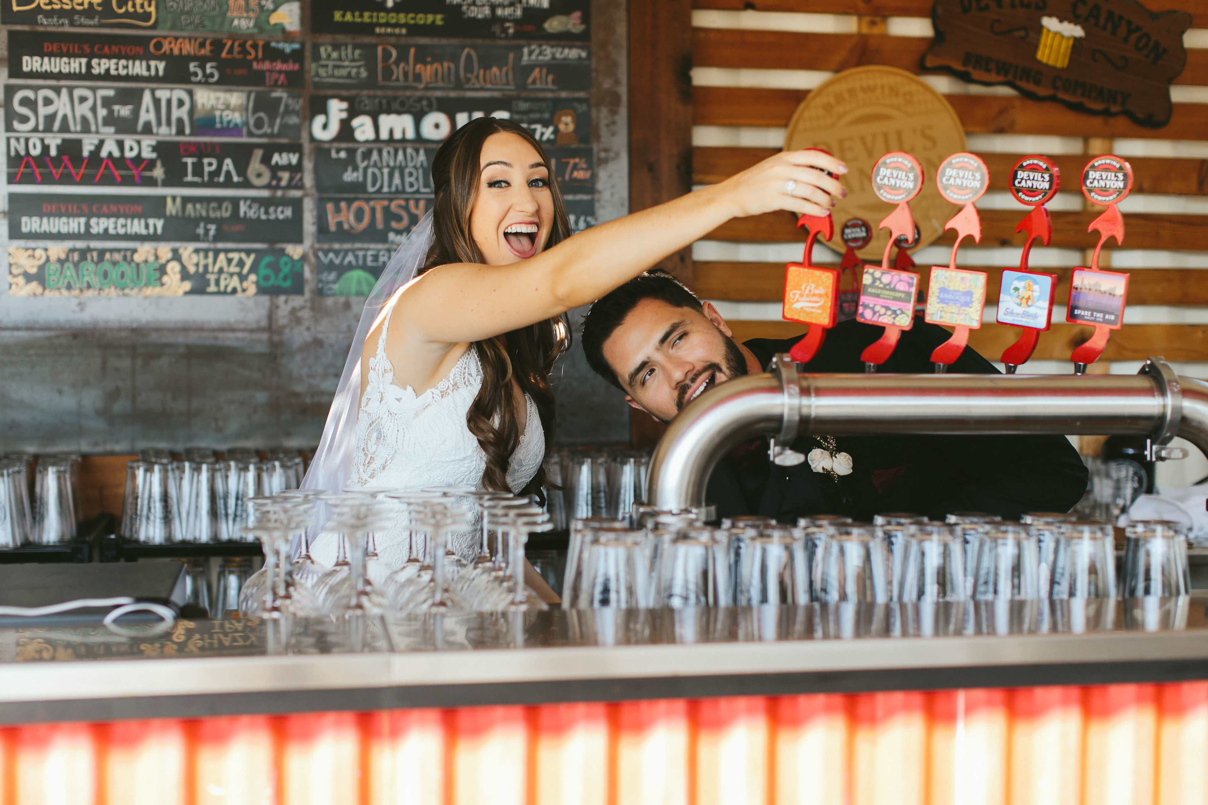 A bride and groom are behind a bar, with the bride playfully holding up drink taps while the groom smiles beside her.