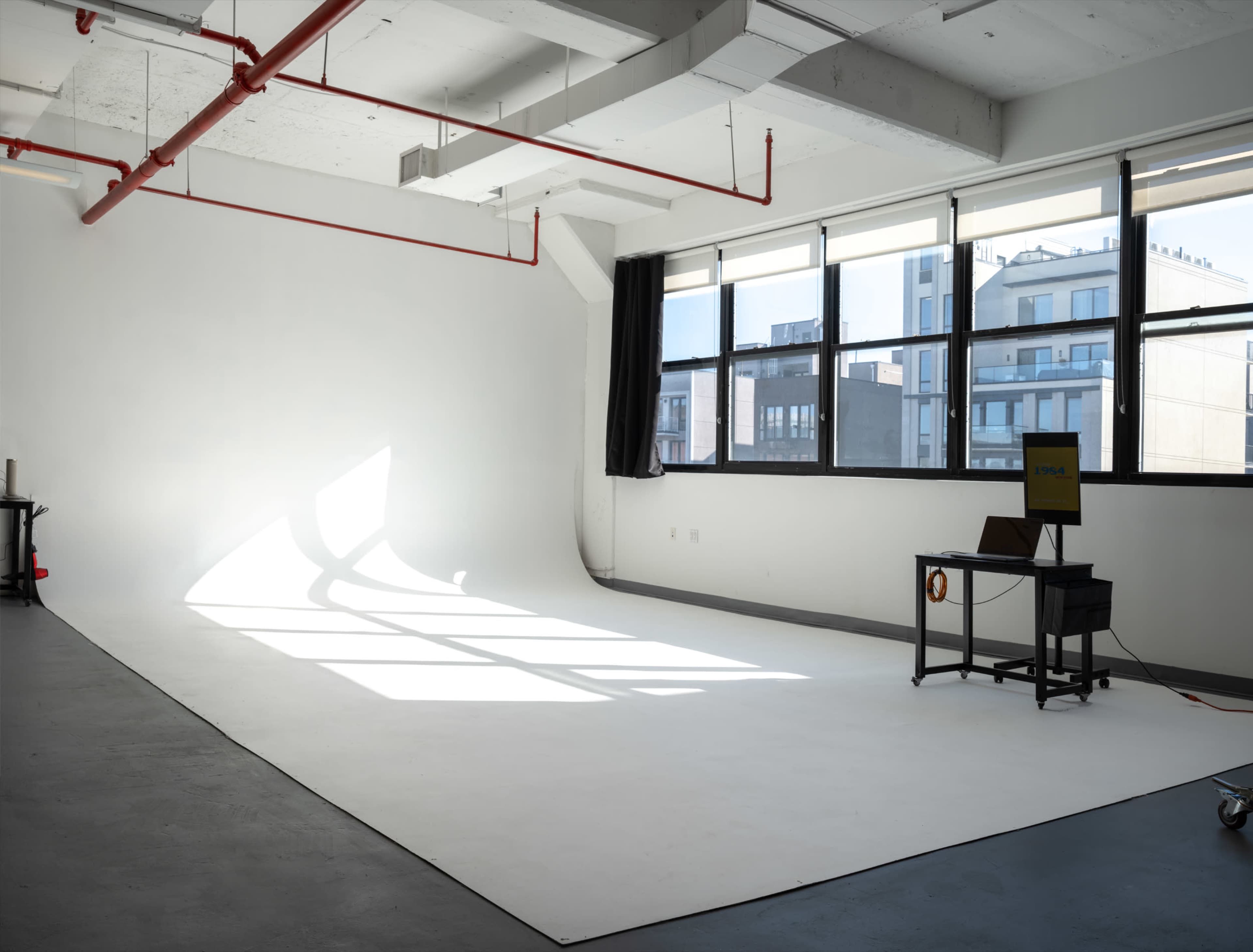 The image shows a spacious photography studio with large windows allowing natural light to illuminate a plain white backdrop on the floor.