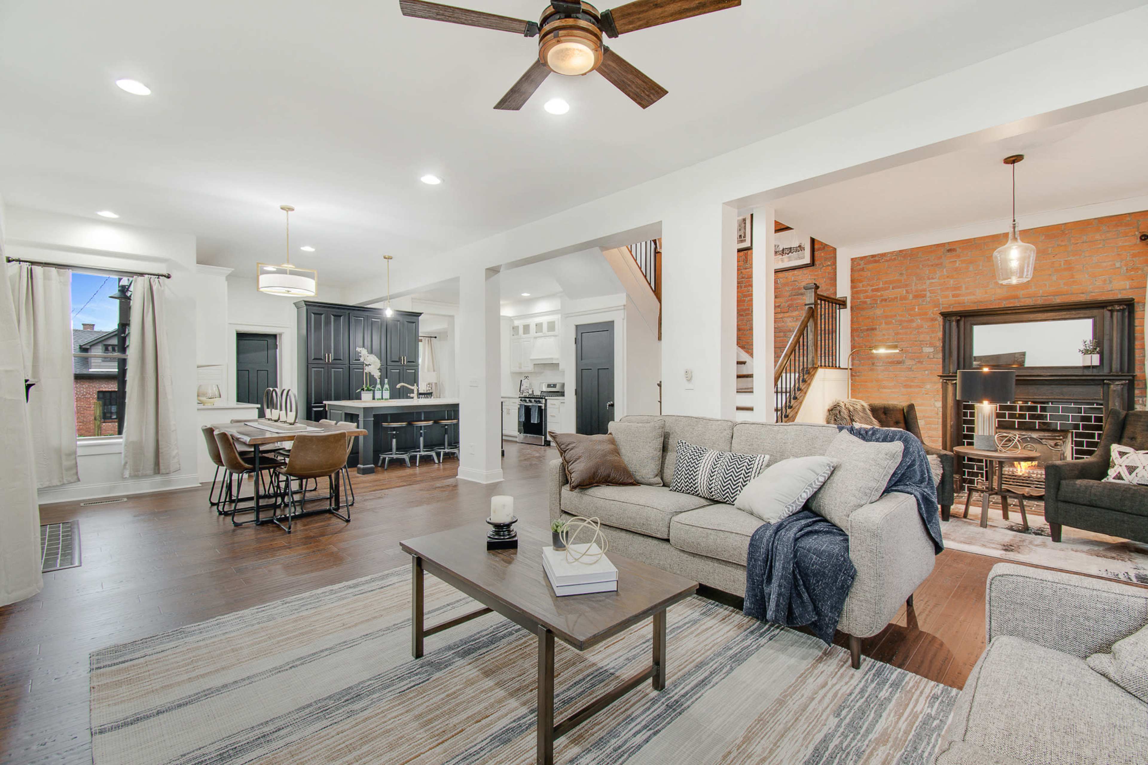 A modern living room with a comfortable sofa, a coffee table, and a view of an adjacent dining area and kitchen, highlighted by warm lighting and a brick accent wall.