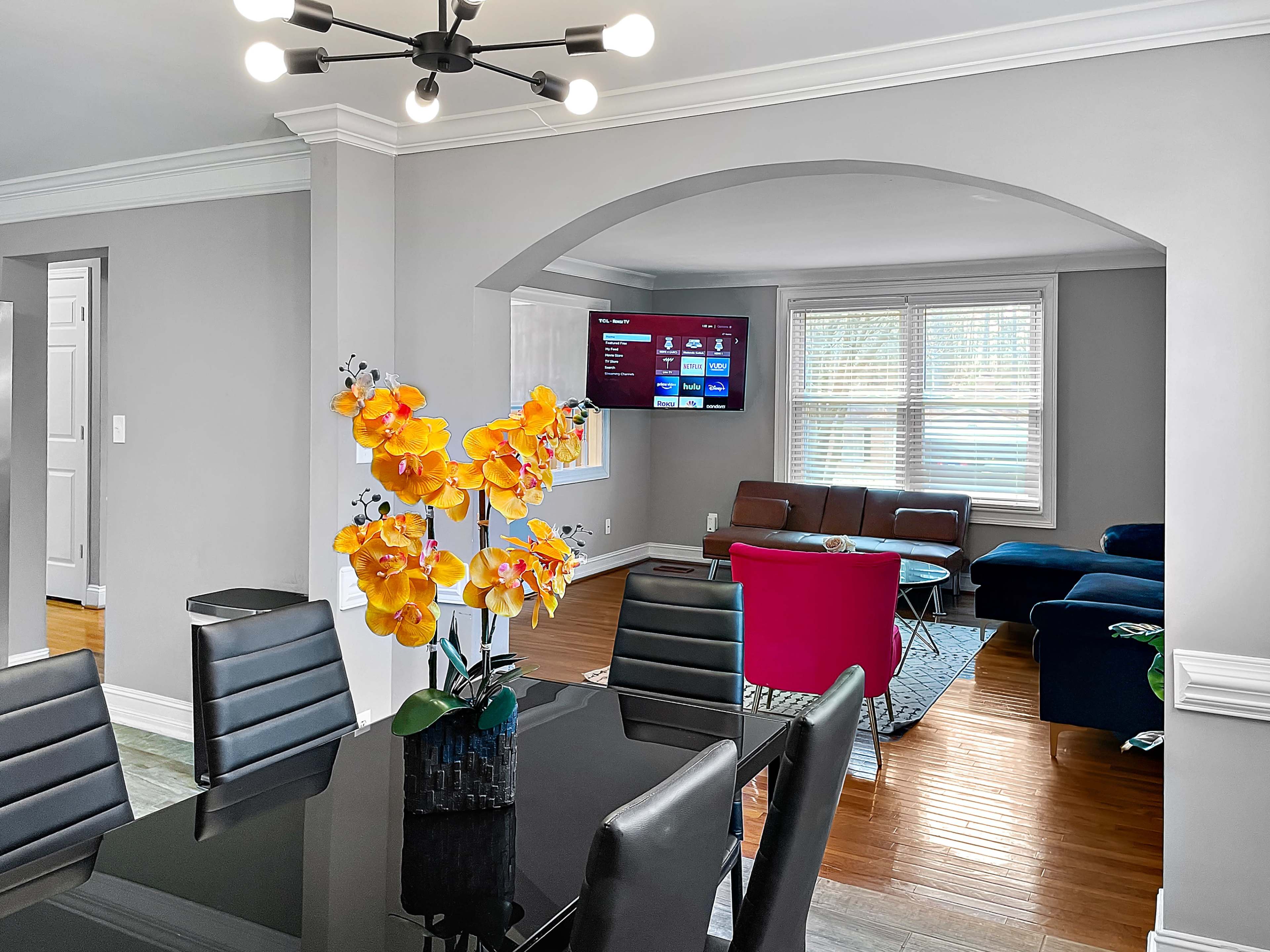 A dining area features a black table with gray chairs and a vase of yellow flowers, adjacent to a living space with a blue sofa and a mounted television on the wall.