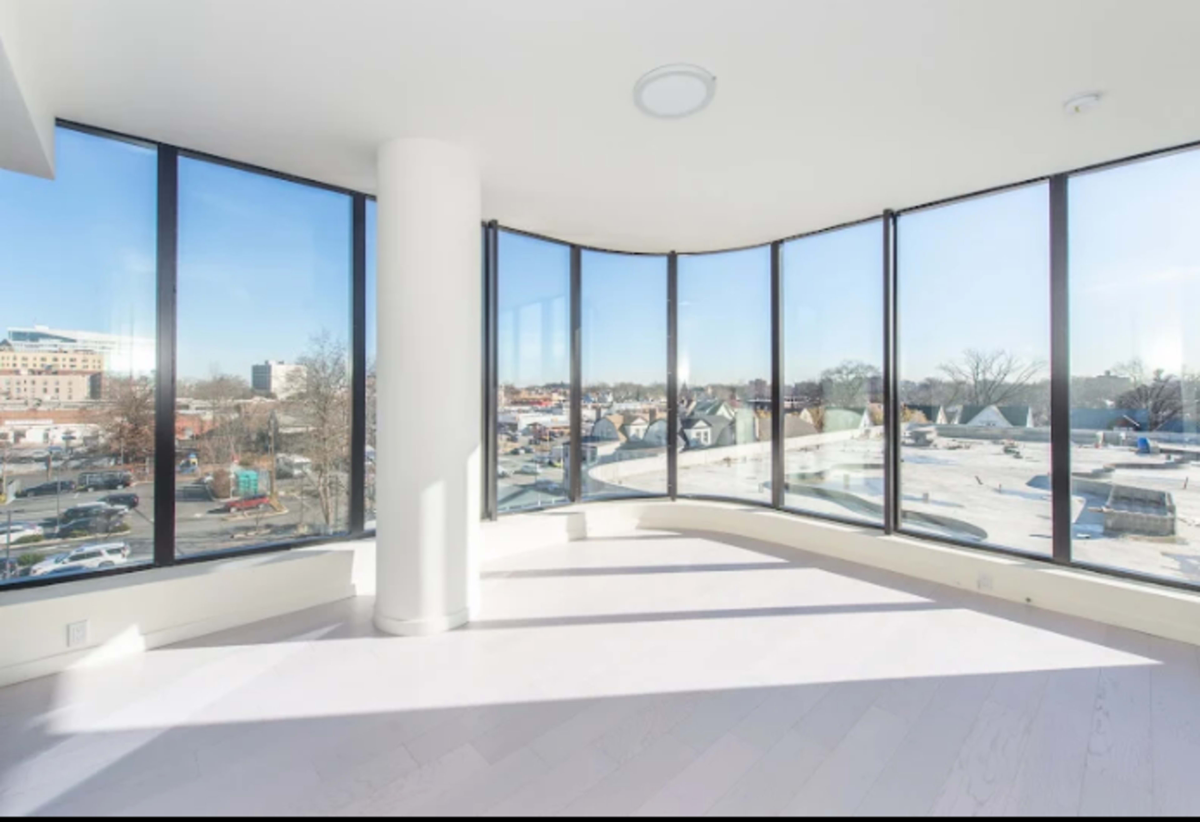 The image shows a modern, spacious room with floor-to-ceiling curved windows offering a view of a construction site and surrounding buildings.
