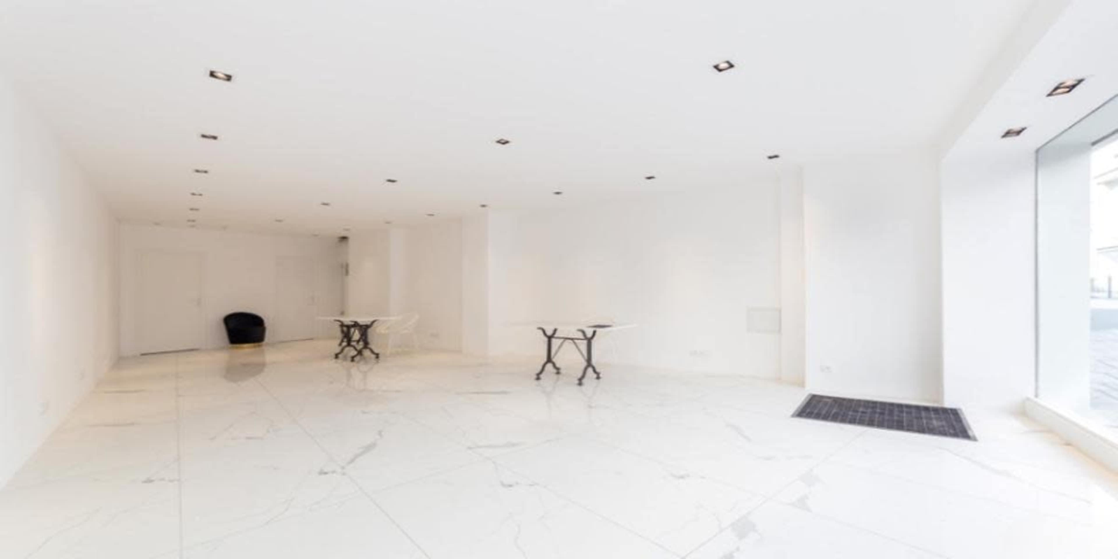 The image shows a bright, empty room with white walls and a glossy marble floor, featuring two small tables and a single black chair in the corner.