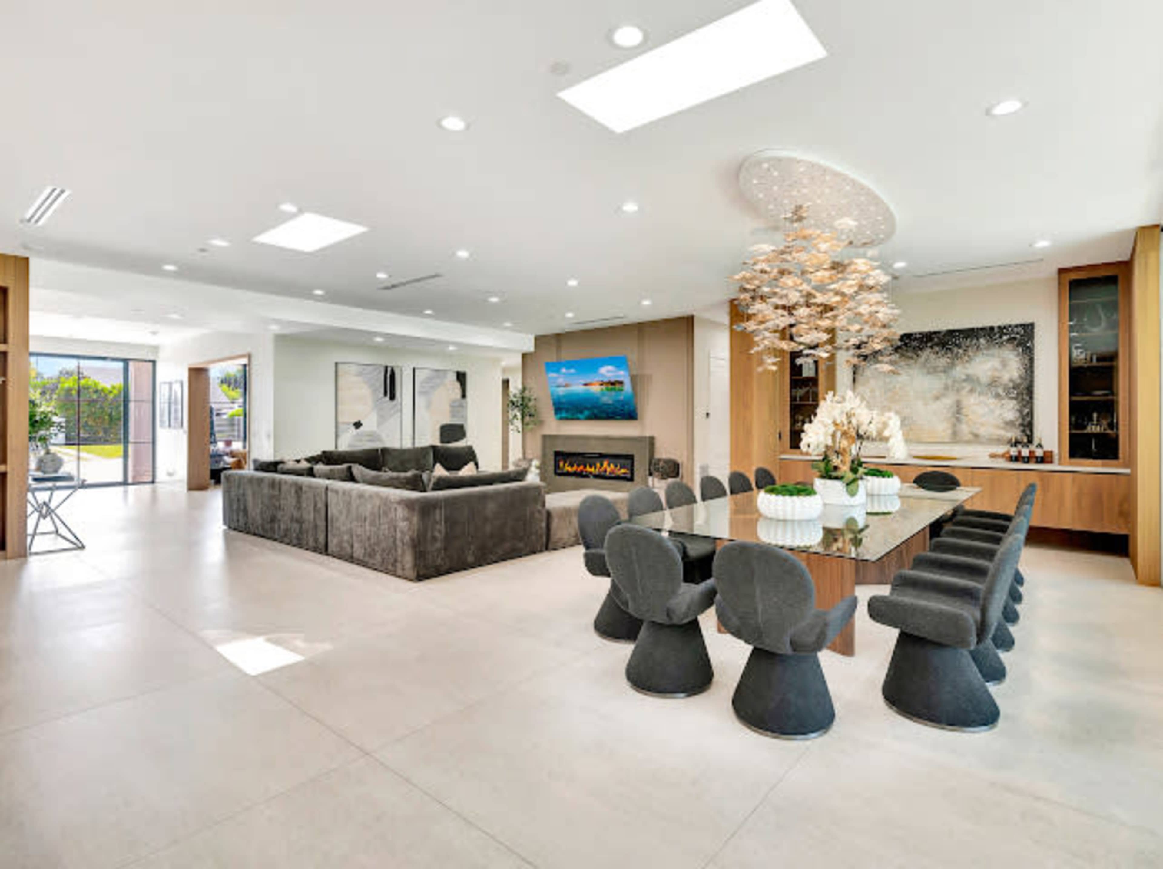The image shows a modern open-concept living and dining area featuring a spacious sectional sofa, a long dining table surrounded by unique chairs, and large windows that provide natural light.