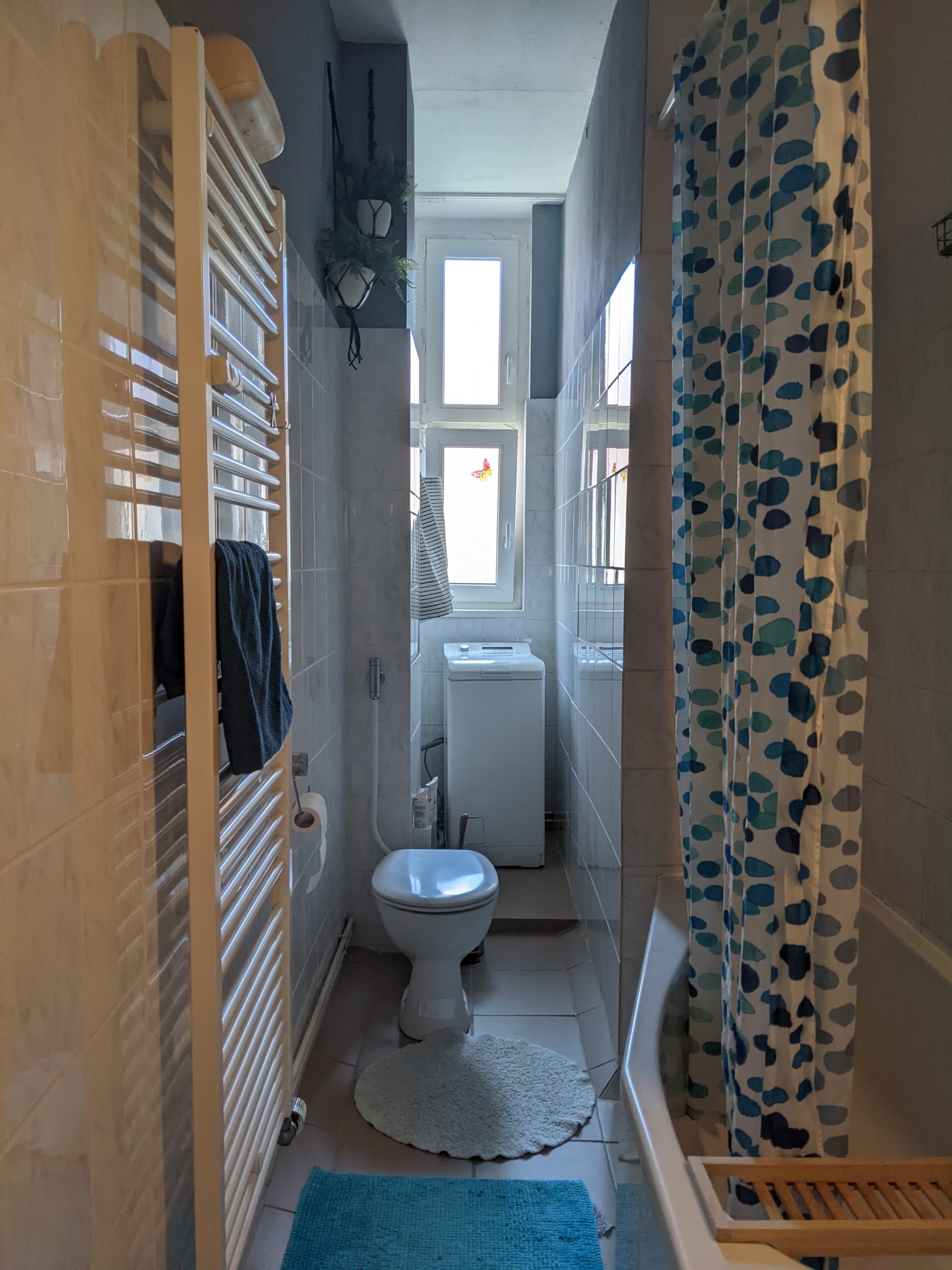 The image shows a narrow bathroom featuring a toilet, a bathtub with a shower curtain, a washing machine, and a heated towel rail.