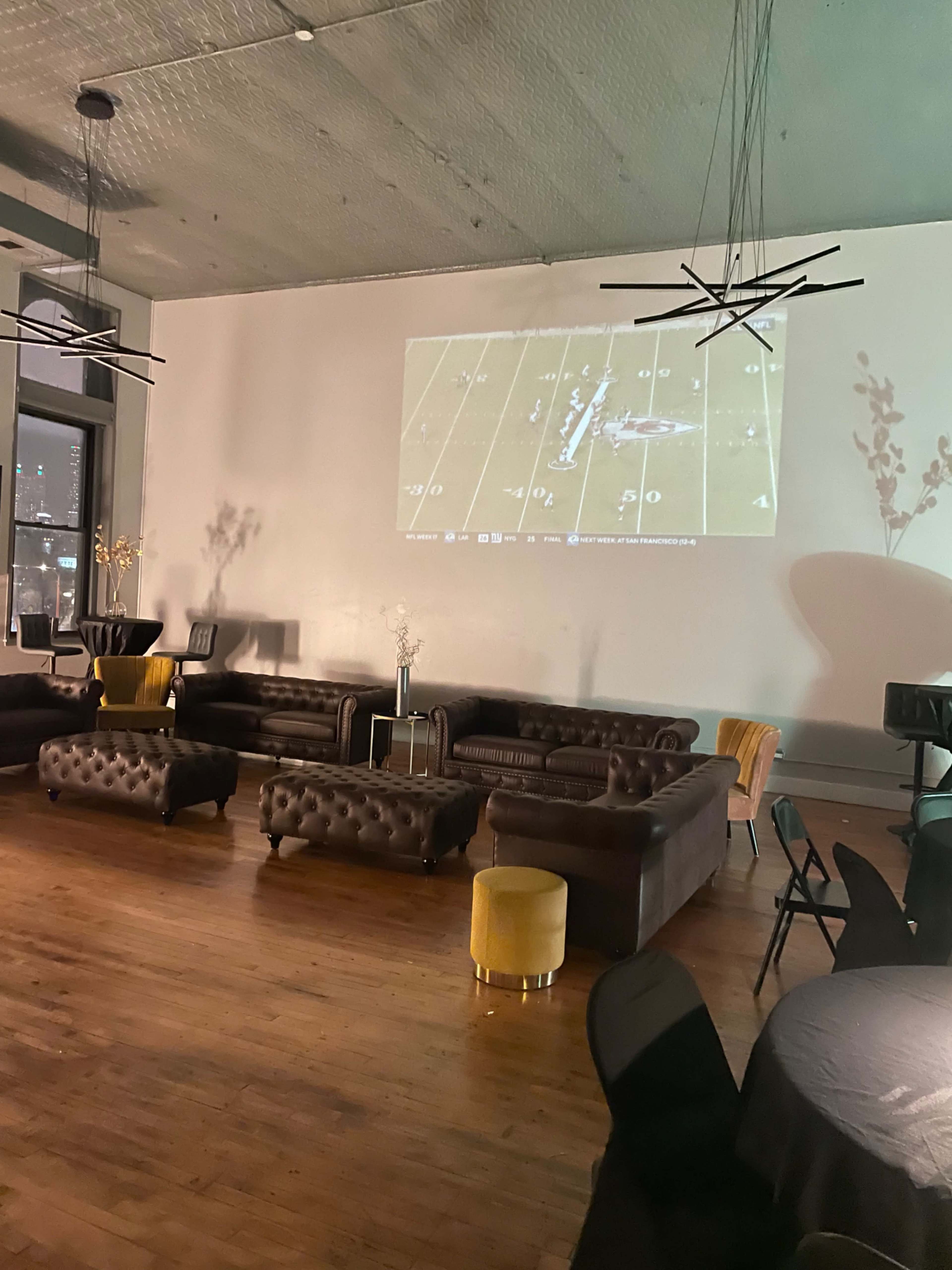The image shows a spacious event room with dark leather couches and chairs, featuring a large screen displaying a sports game on the wall.