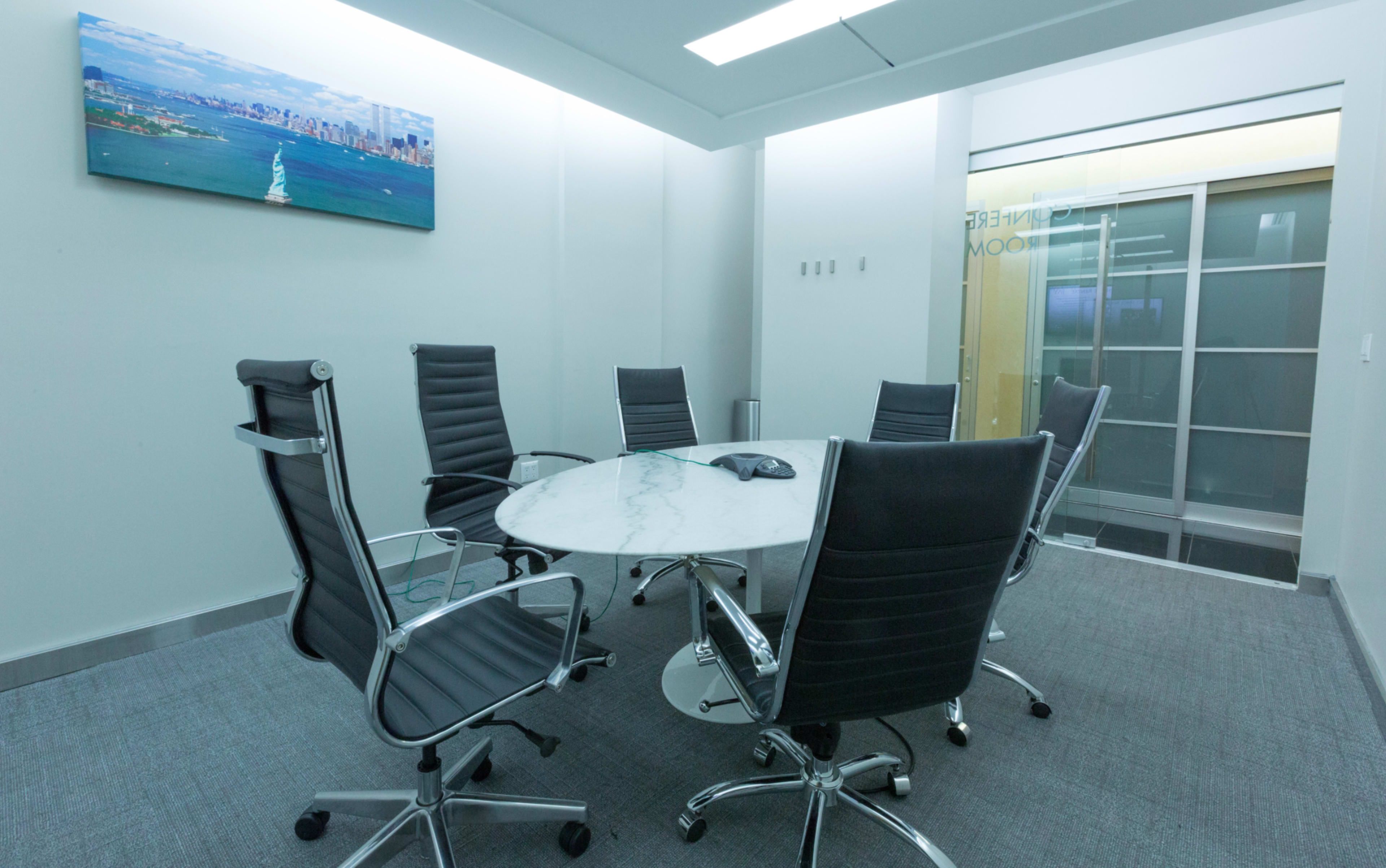 A modern conference room features a circular marble table surrounded by six black leather office chairs, with a large window displaying a cityscape and a photograph of the Statue of Liberty on the wall.