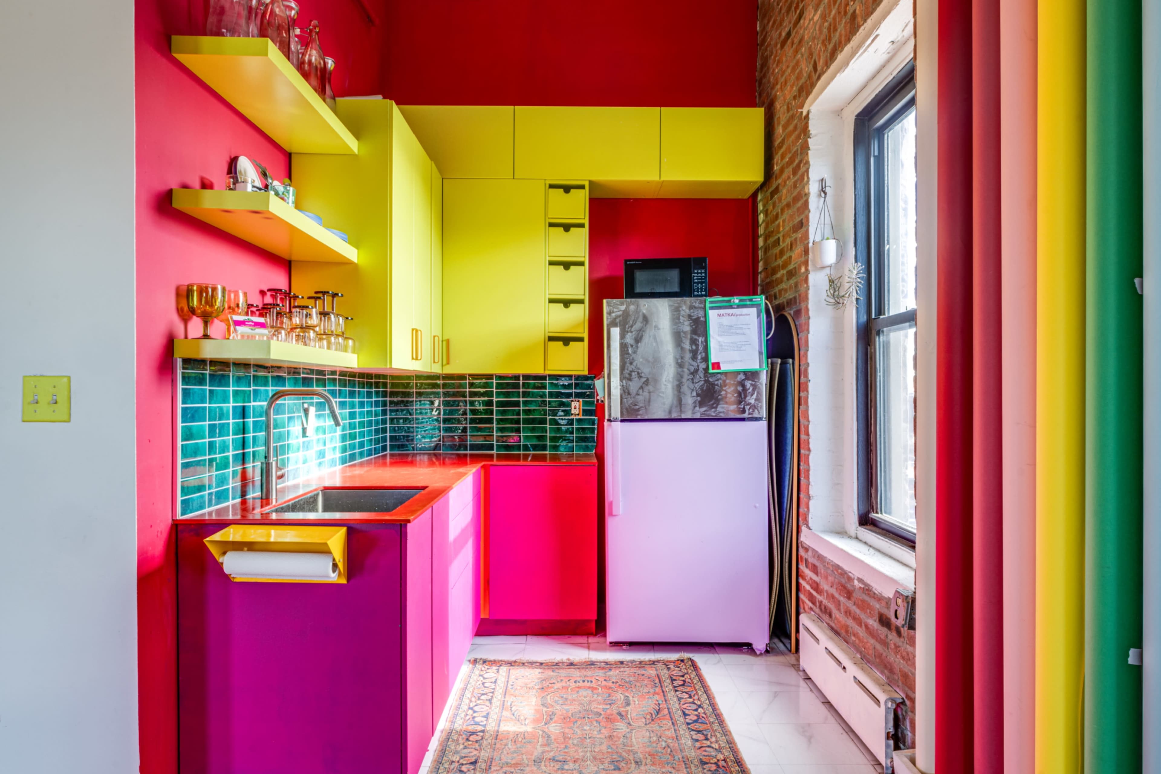 The image shows a brightly colored kitchen with yellow and pink cabinets, a green tiled backsplash, and a white refrigerator against a red wall.