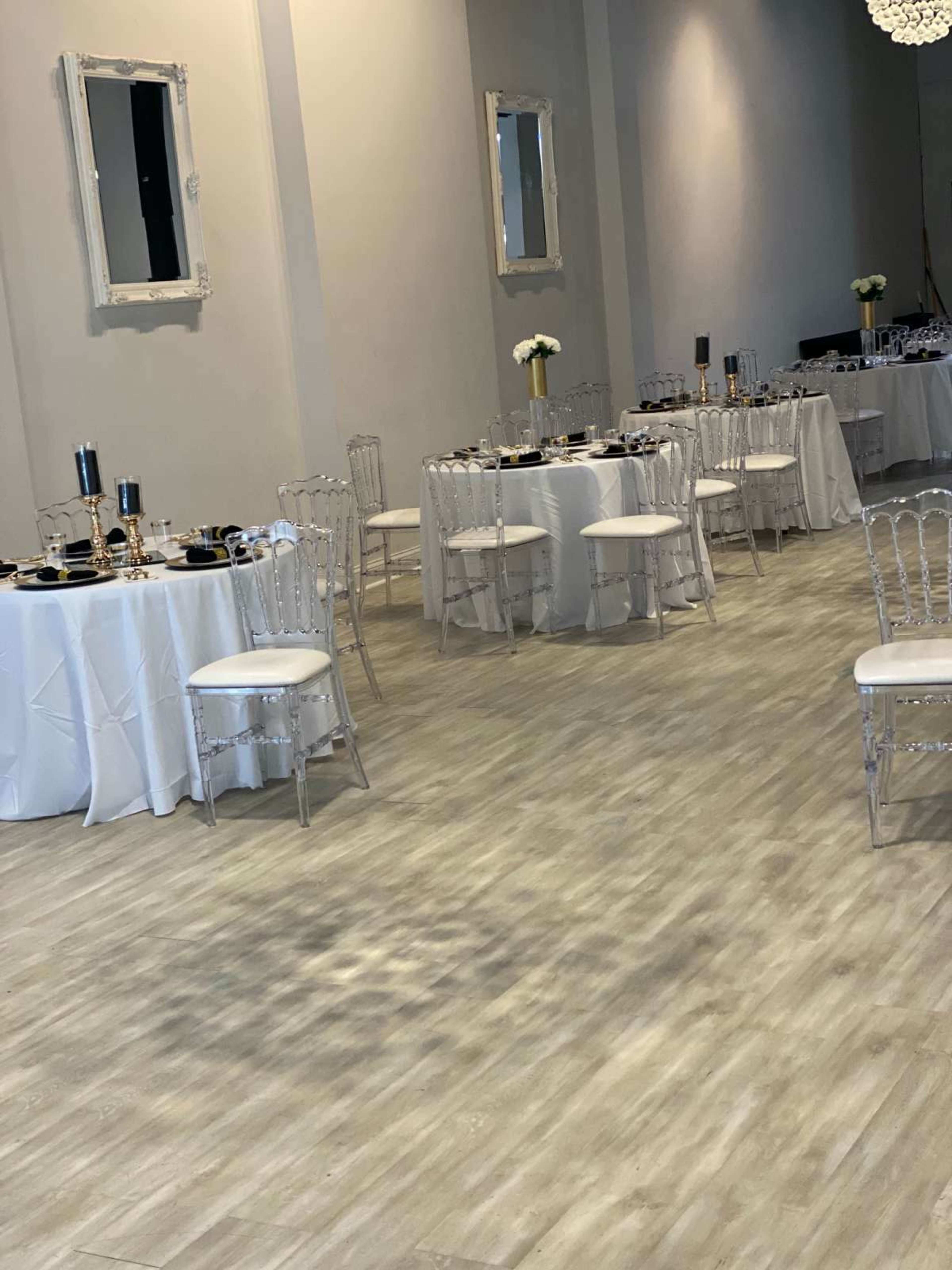 The image shows a spacious event room with several elegantly set tables, each covered in white tablecloths and surrounded by clear acrylic chairs.