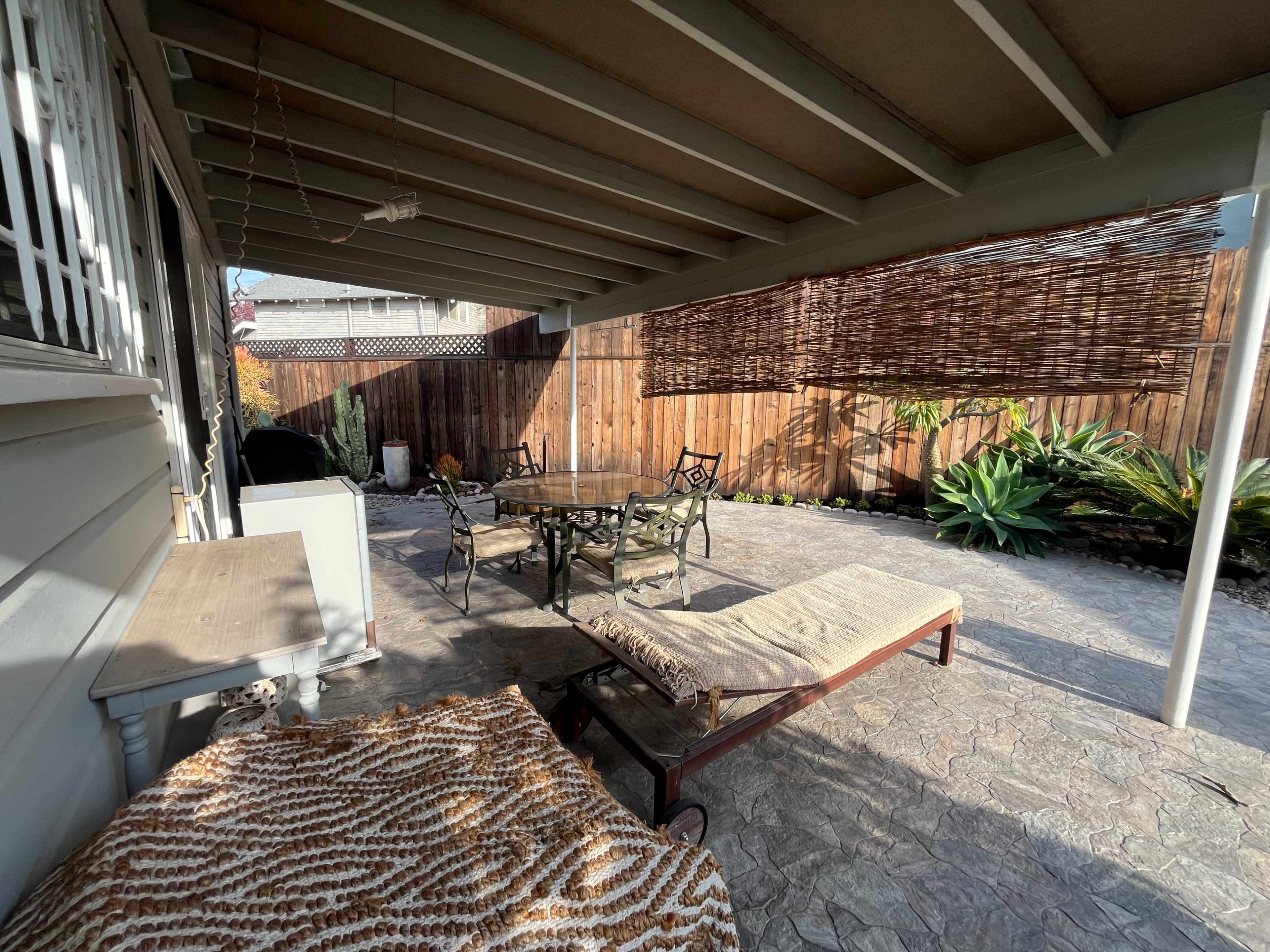 The image shows a patio area with a seating arrangement, including a table and chairs, surrounded by plants and a wooden fence.