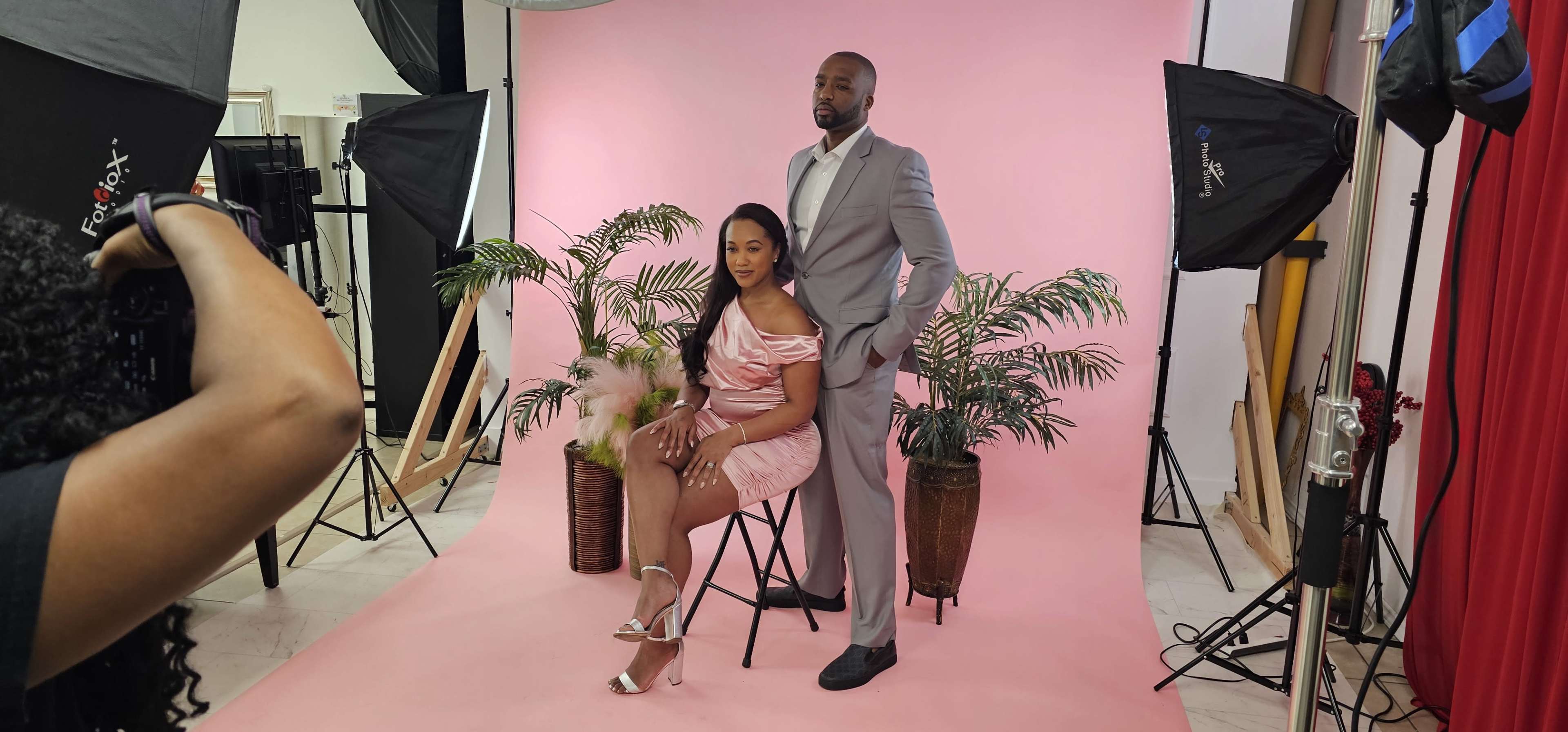 A man in a gray suit stands behind a woman in a pink dress seated on a chair, with palm plants and studio lights arranged around them against a pink backdrop.