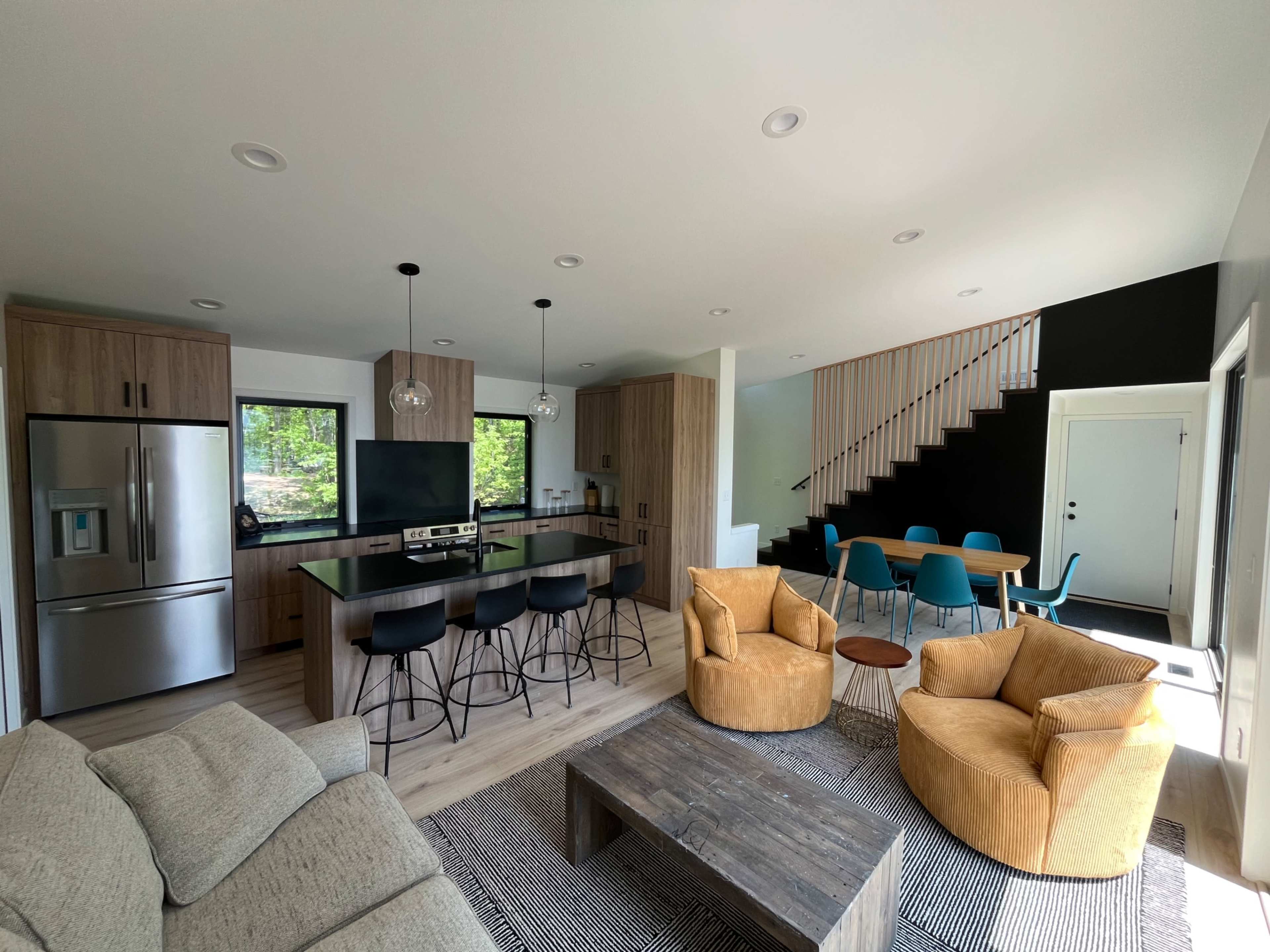 The image shows a modern open-concept living space featuring a kitchen with a large island, dining area with teal chairs, and a cozy seating area with two large armchairs.