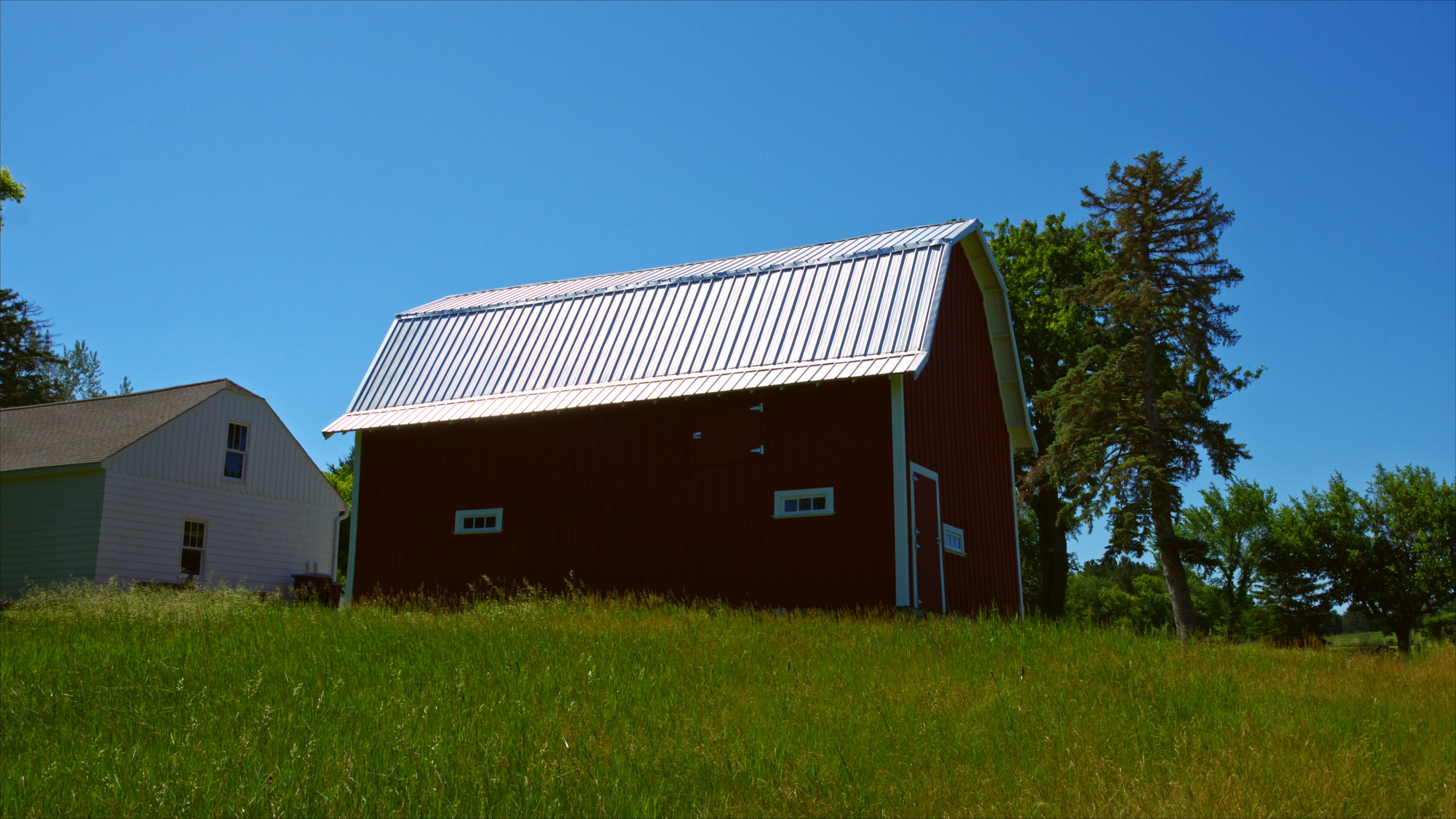 A red barn with a metal roof stands on a grassy field, next to a white building under a clear blue sky.