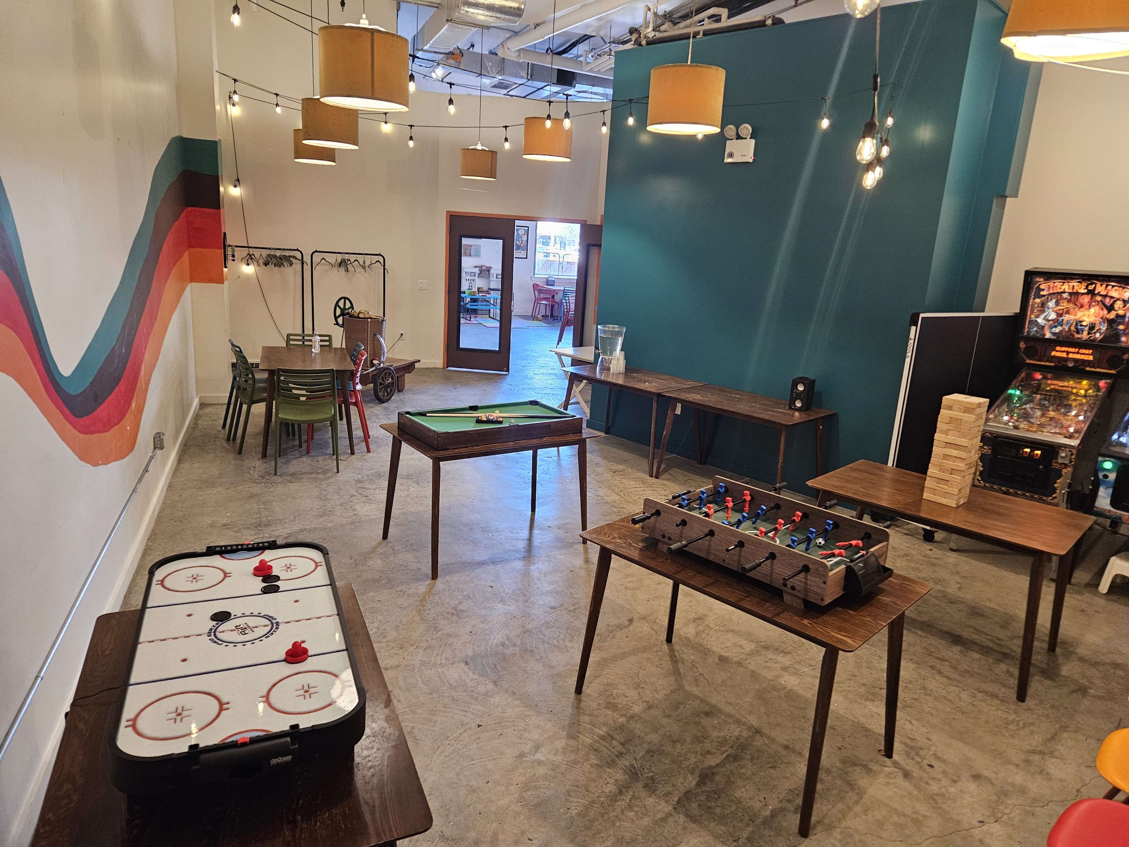 The image shows a recreational room with various gaming tables, including an air hockey table, a foosball table, and a stack of Jenga blocks, all under warm hanging lights.