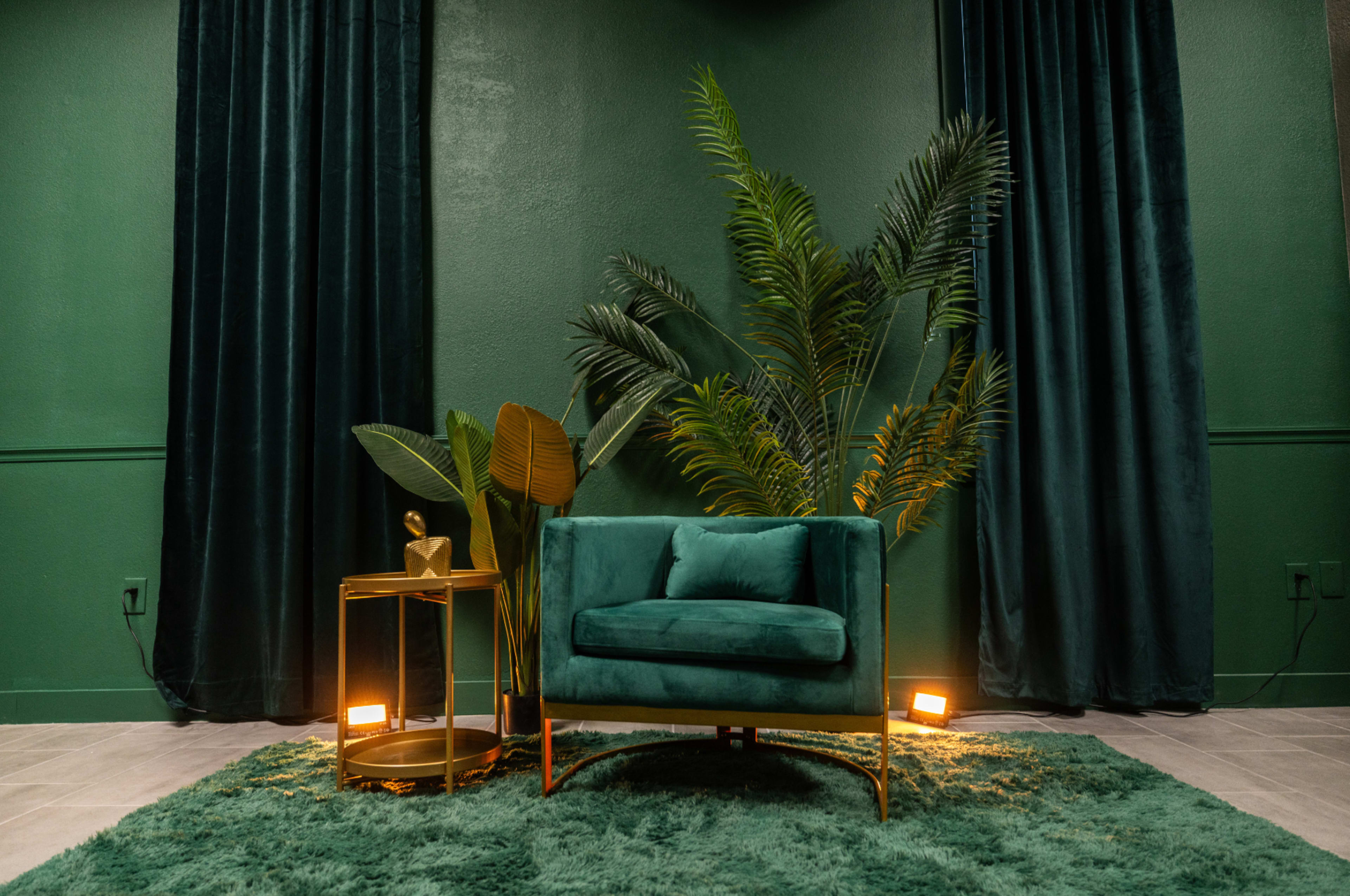 A teal armchair sits next to a gold side table, surrounded by large green plants and dark green curtains on a textured rug.