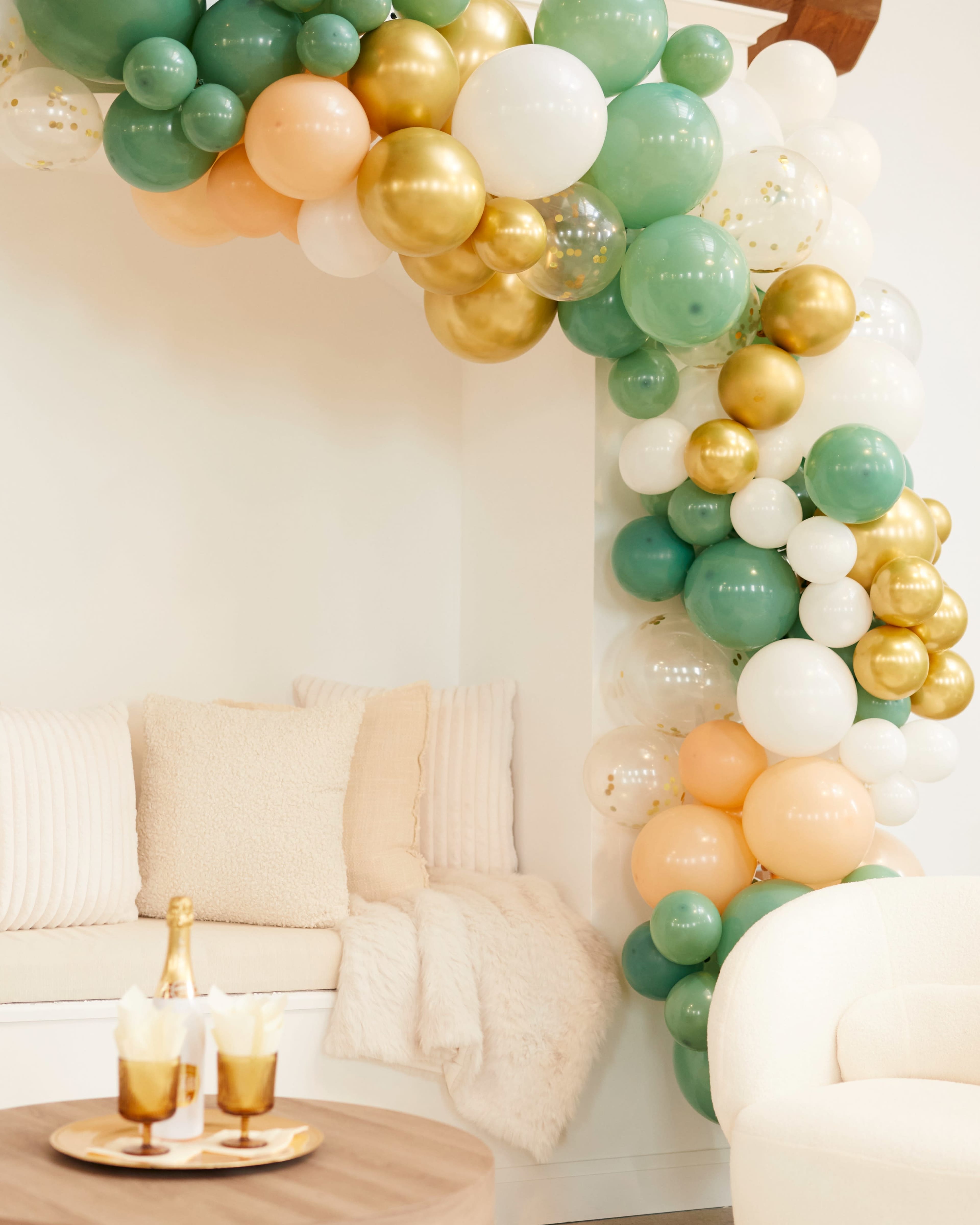 A decorative arch made of colorful balloons in shades of green, peach, gold, and white is positioned beside a cozy seating area.