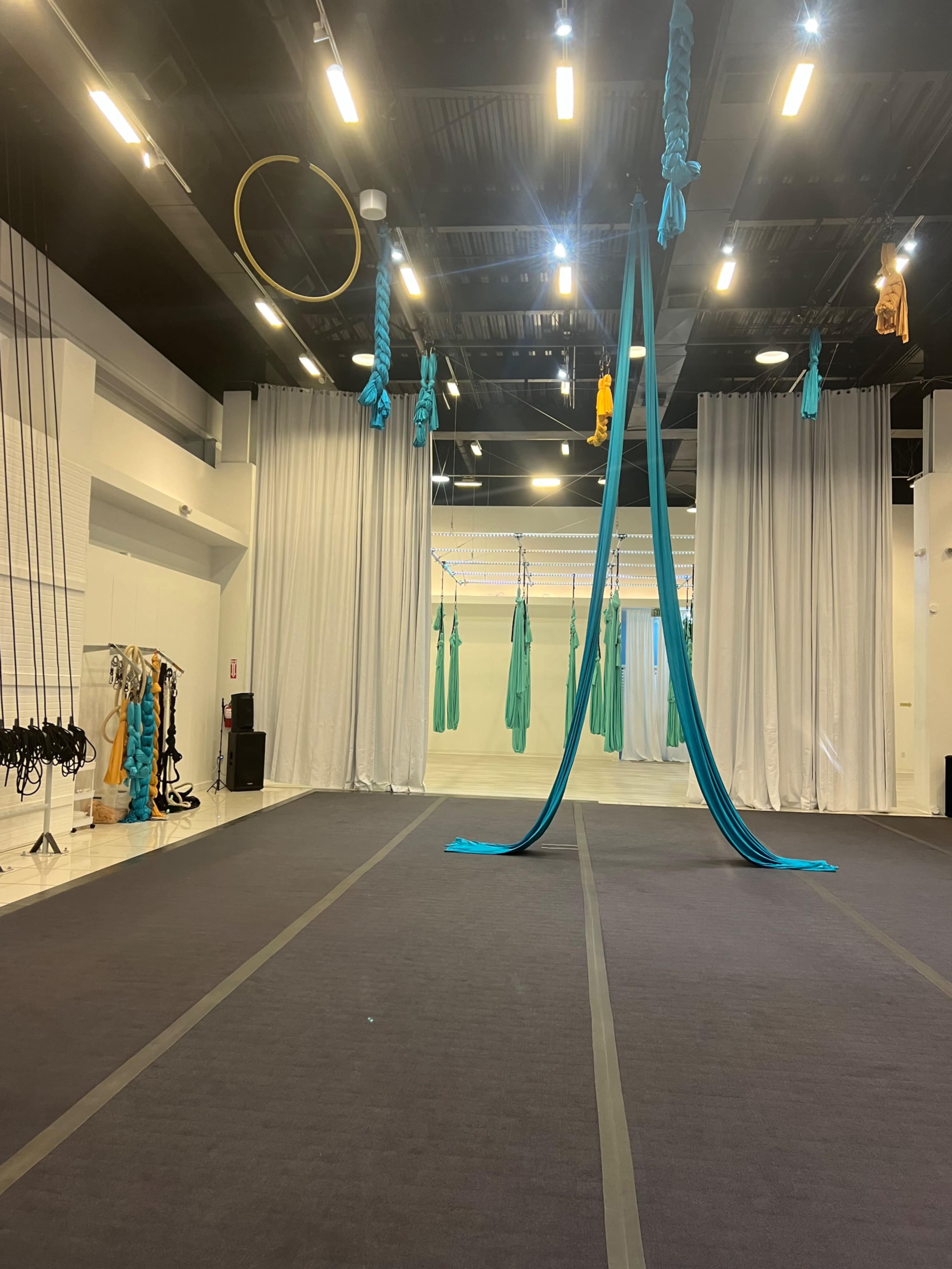 An aerial yoga studio with hanging silks, hoops, and a black floor mat surrounded by white curtains.