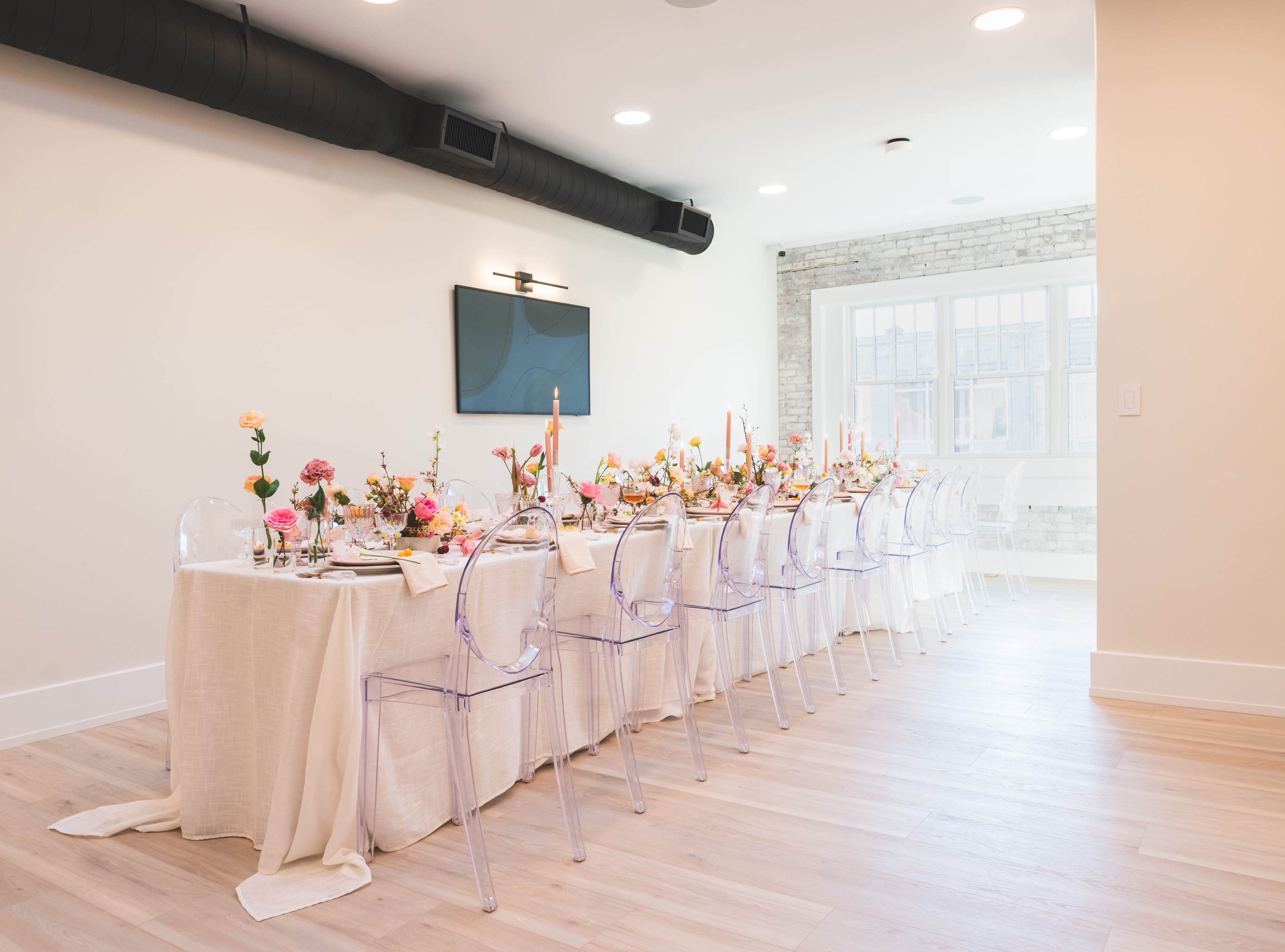 A long dining table decorated with flowers and candles sits under a modern light fixture in a bright room with a large window.