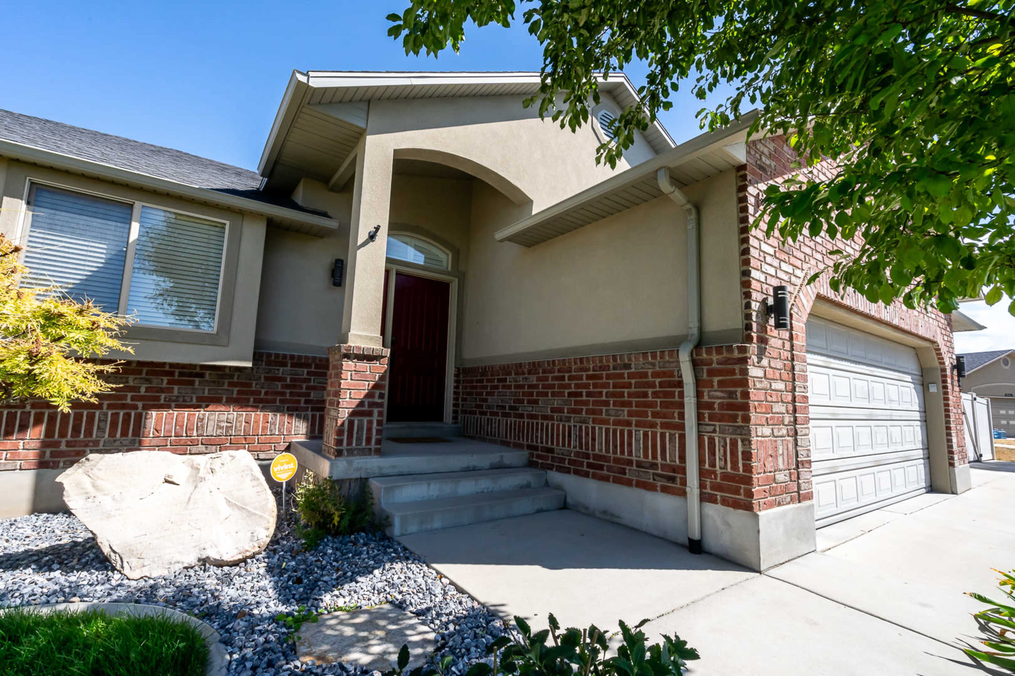 The image shows the entrance of a single-story house featuring a brick and stucco exterior, with a landscaped yard and a paved walkway leading to the door.