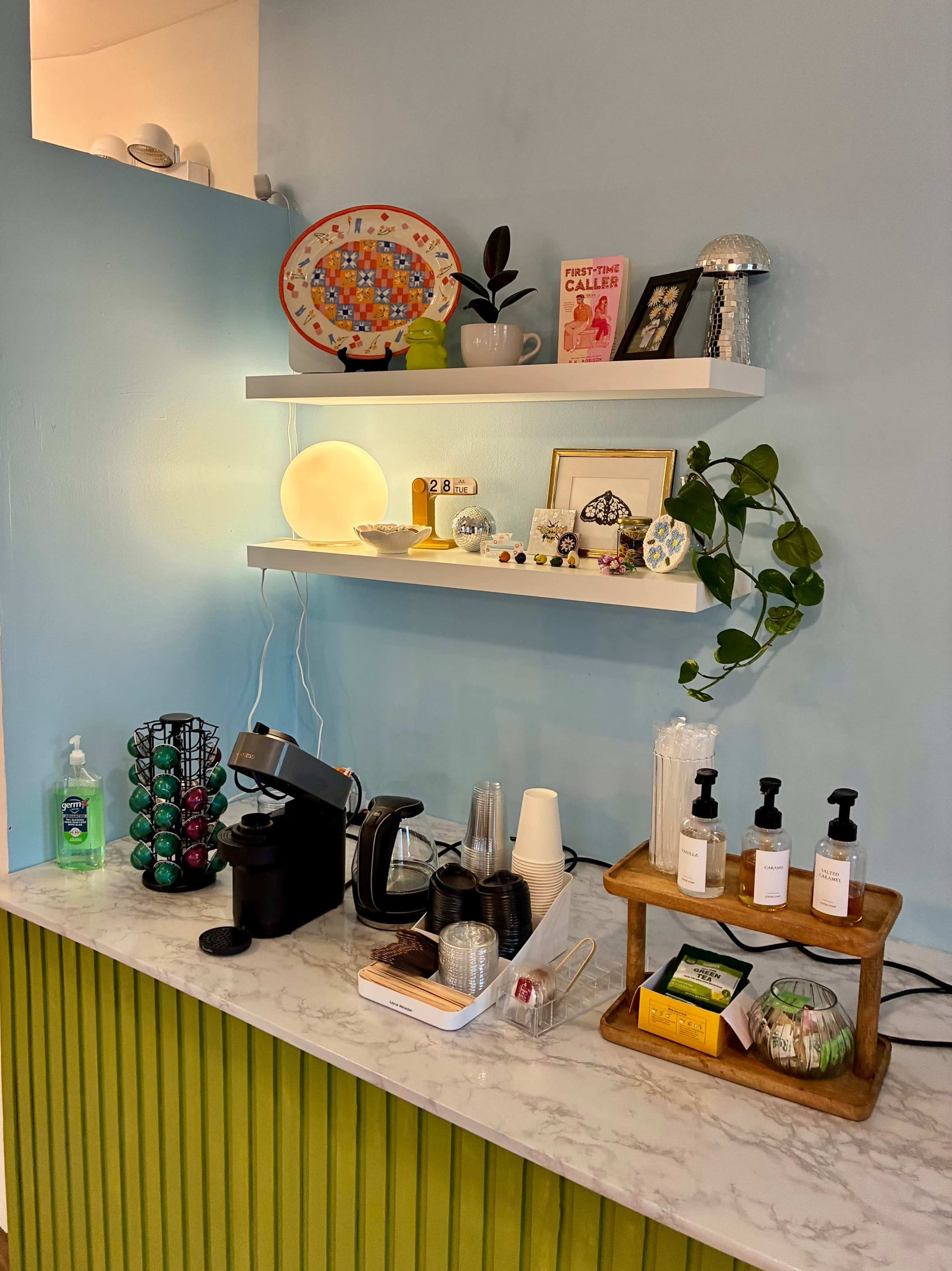The image shows a small coffee station with a coffee maker, various coffee pods, cups, and toiletries on a marble counter beneath a light blue wall with decorative shelves.