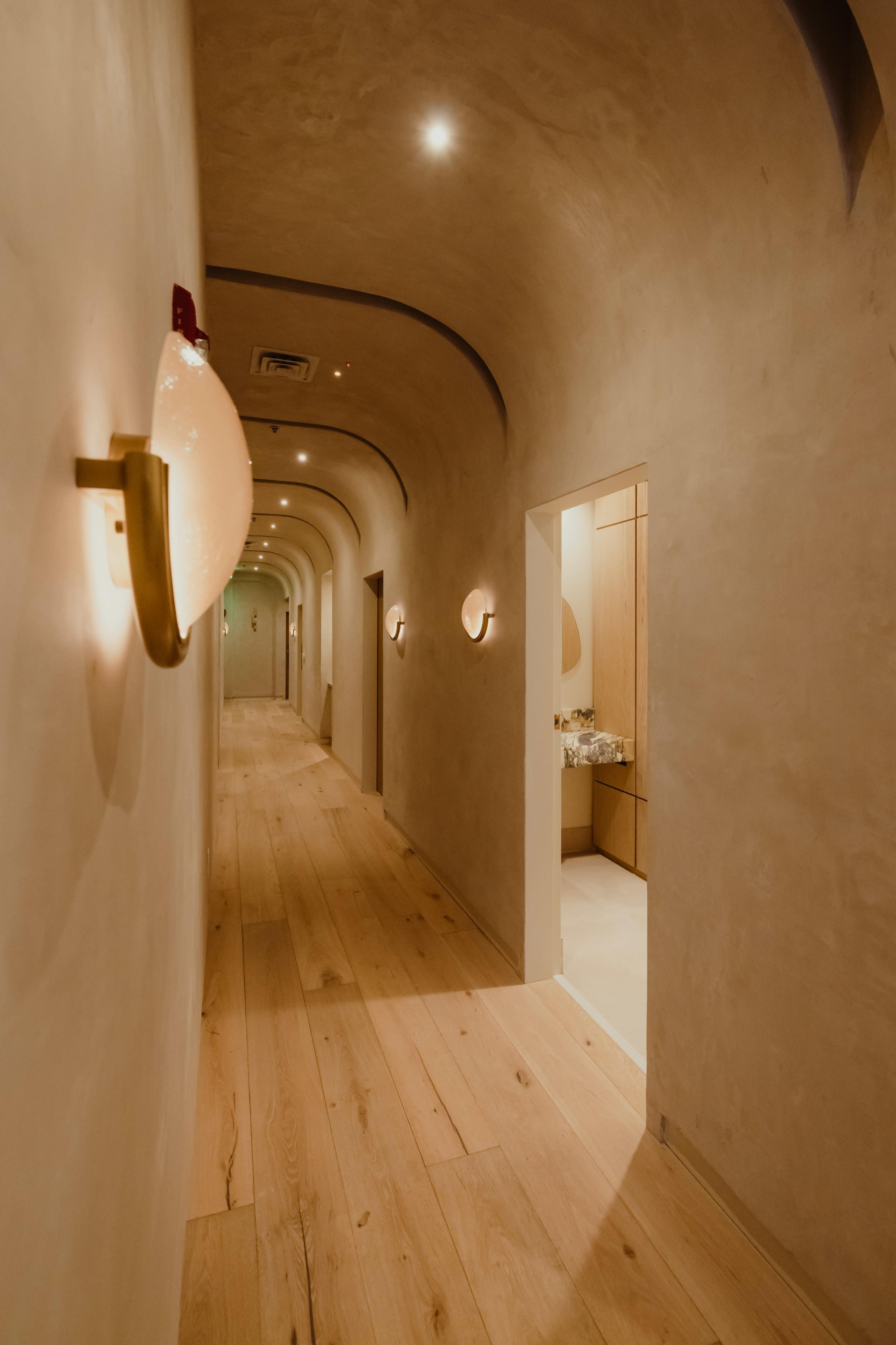 The image depicts a softly lit, arched hallway with wooden flooring and light fixtures on the walls.