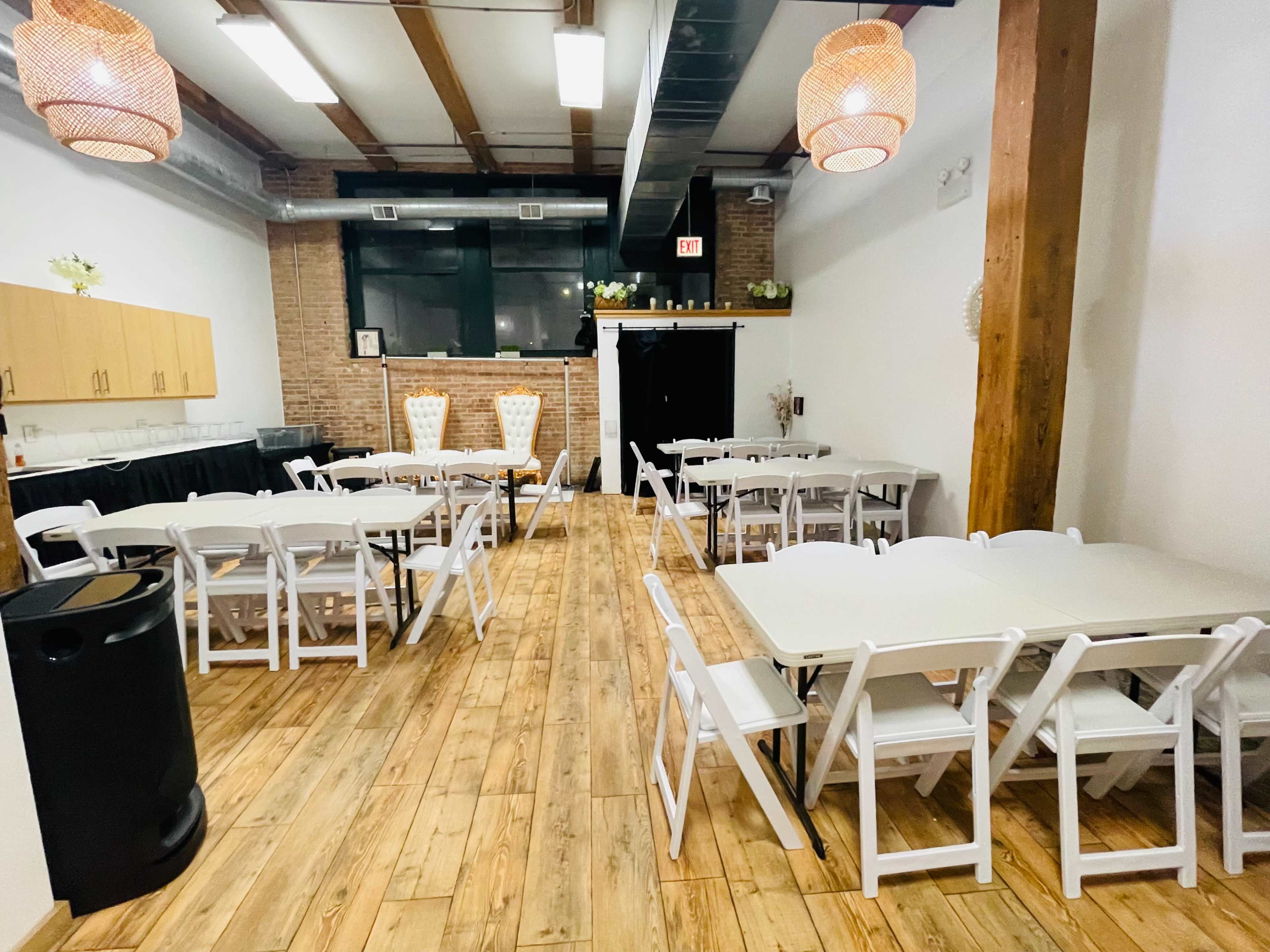 The room features several arranged tables with white chairs, warm wooden flooring, exposed brick walls, and pendant lights.