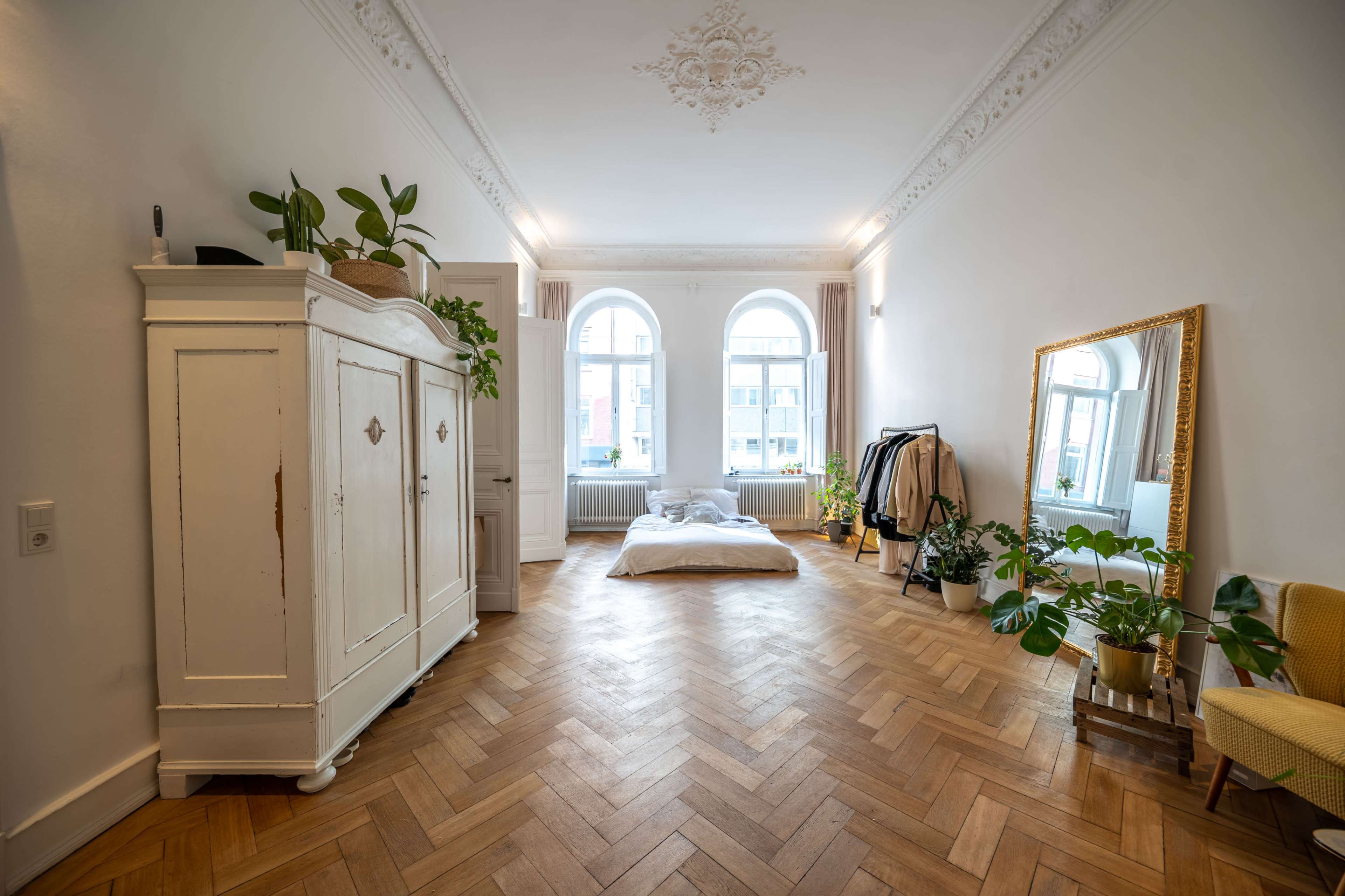 The room features a bed with white bedding, a large mirror, wooden flooring, and several plants arranged around the space.