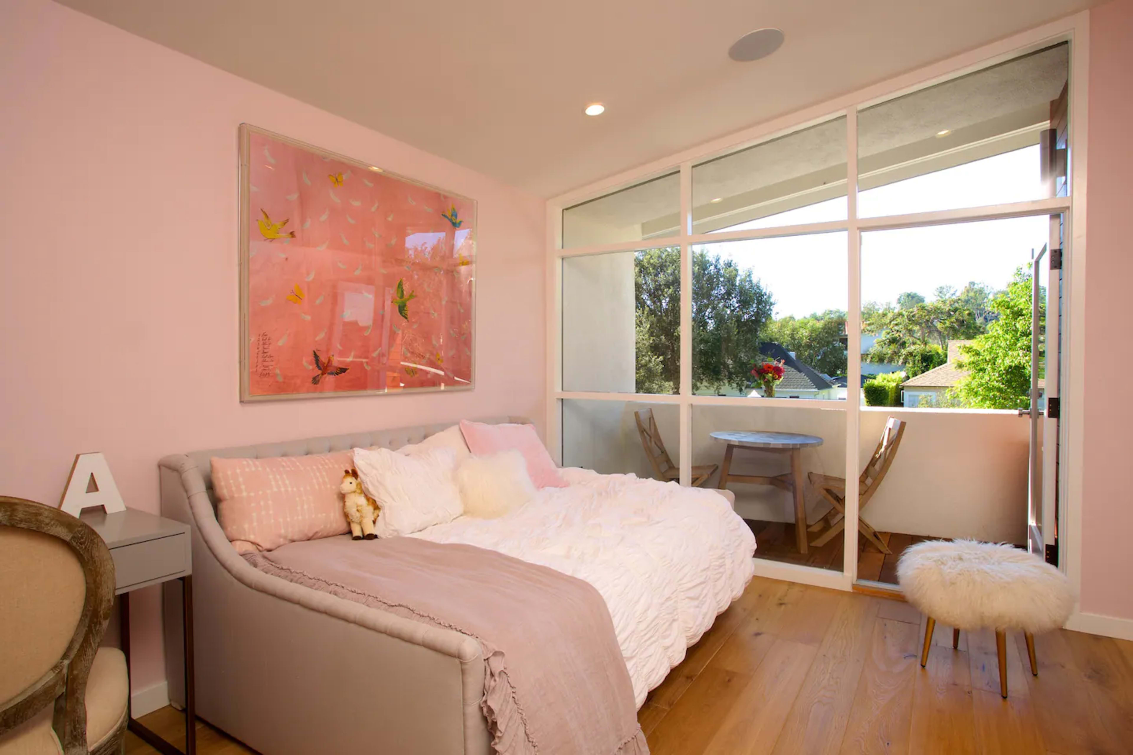 A light pink bedroom features a small bed with white bedding, a nearby glass door leading to a balcony, and a round table set with a vase of flowers.