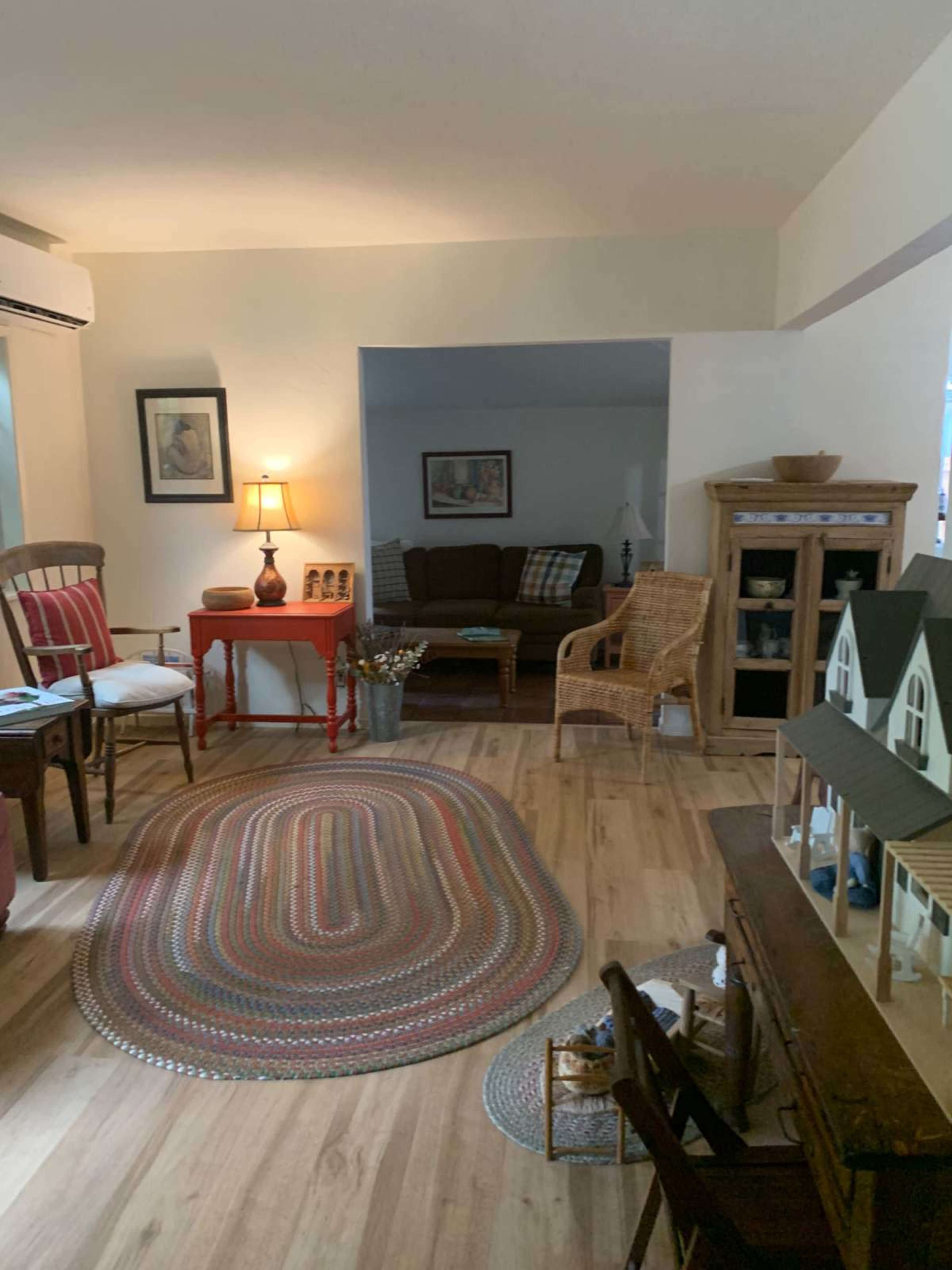 The image shows a cozy living room with wooden flooring, a braided area rug, a lamp, a sofa, and various pieces of furniture arranged around the space.