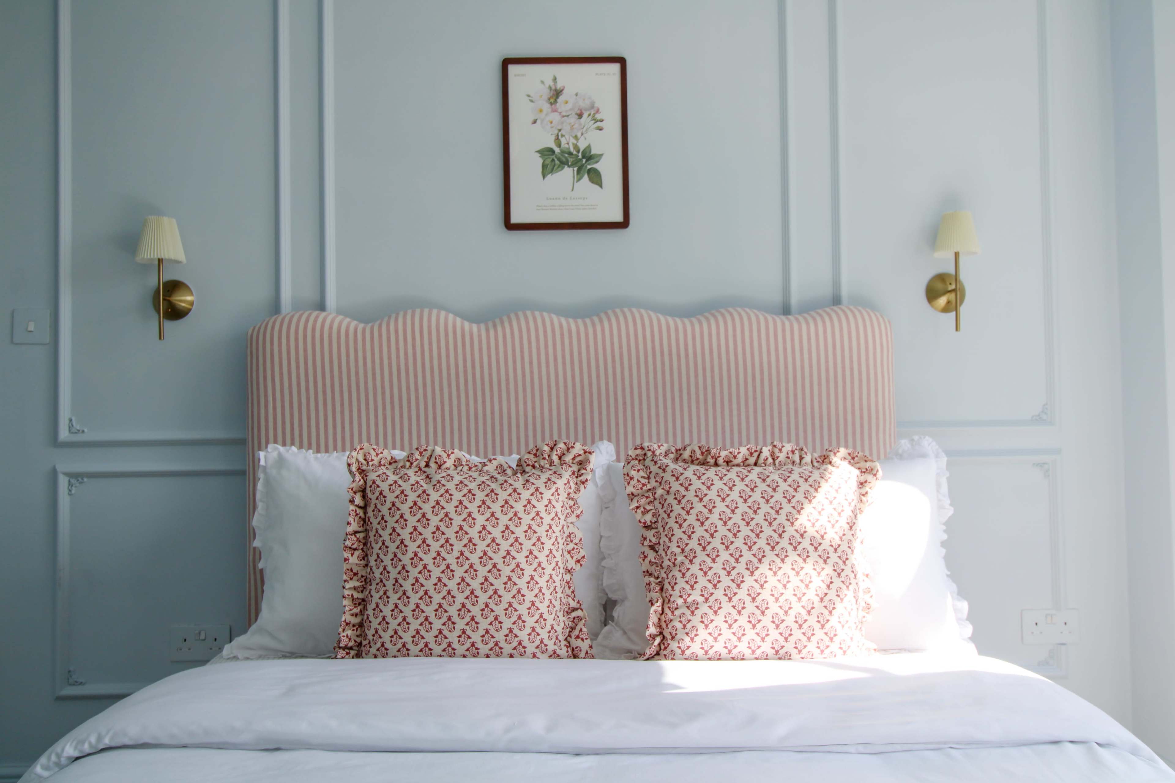 The image shows a neatly made bed with a pink striped headboard, decorative pillows, and a framed botanical print on a light blue wall.