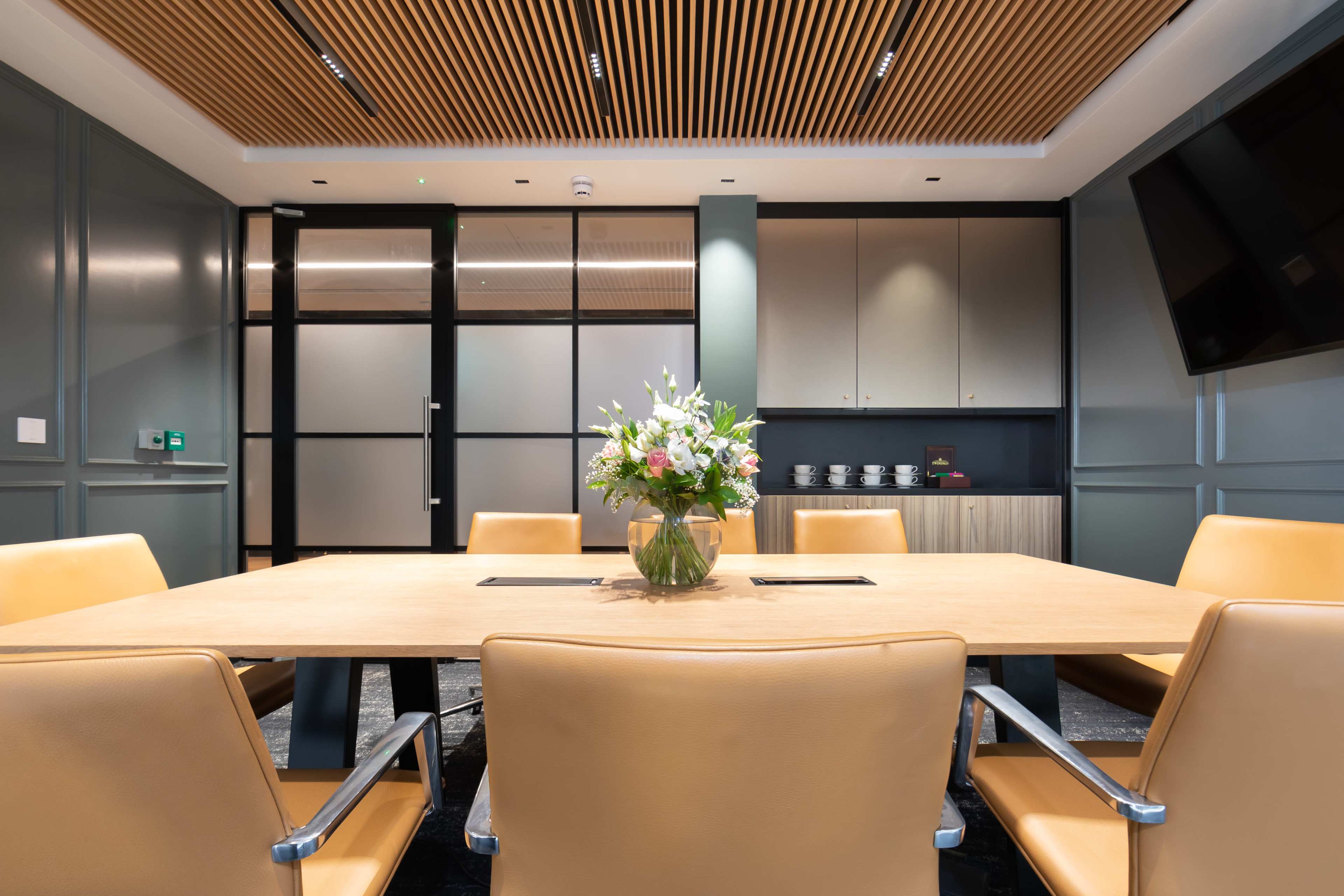 A modern conference room features a large wooden table surrounded by leather chairs, with a floral centerpiece and cabinets in the background.