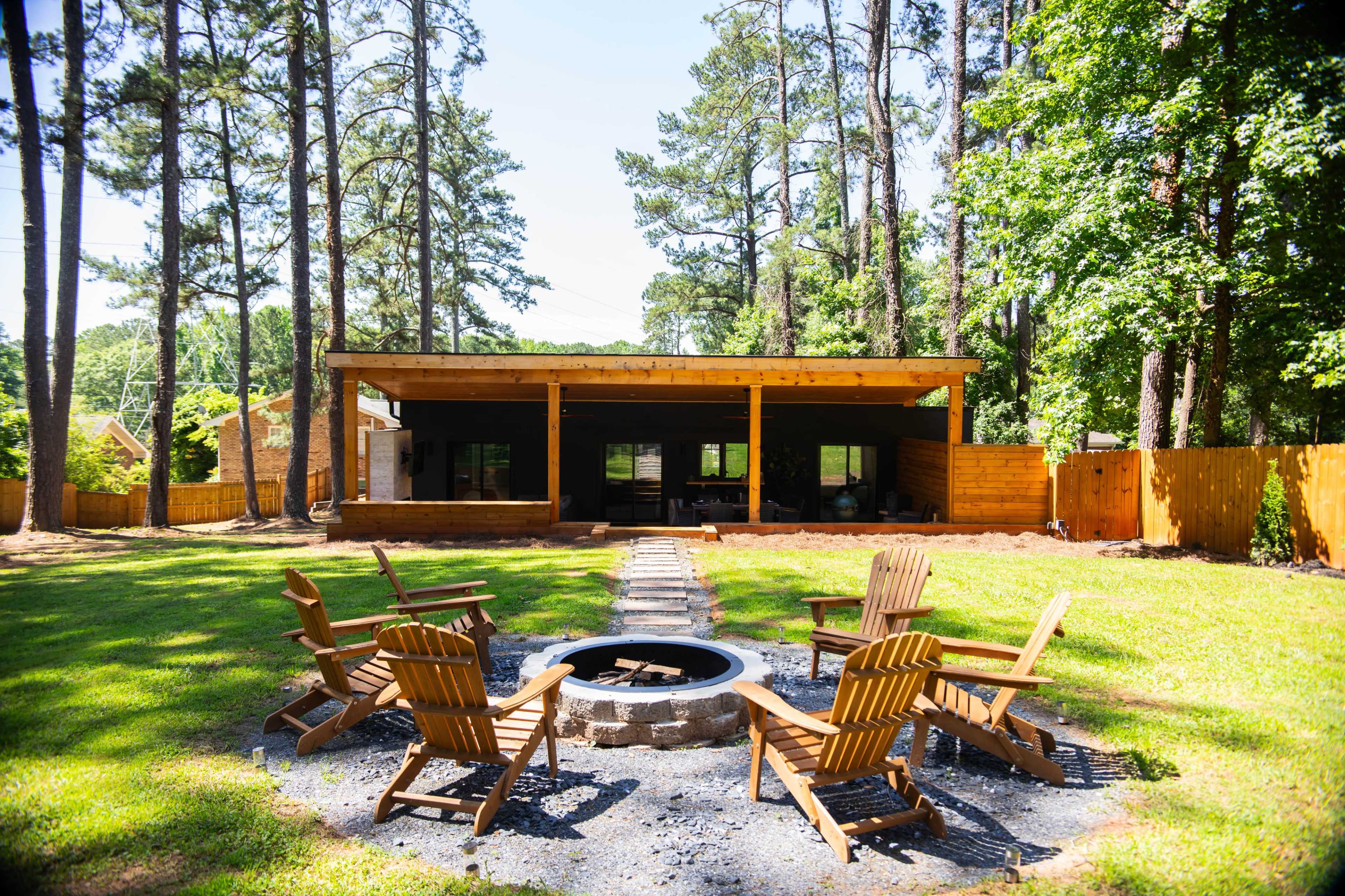 A circular arrangement of wooden chairs surrounds a stone fire pit in a green yard adjacent to a modern wooden structure and tall trees.